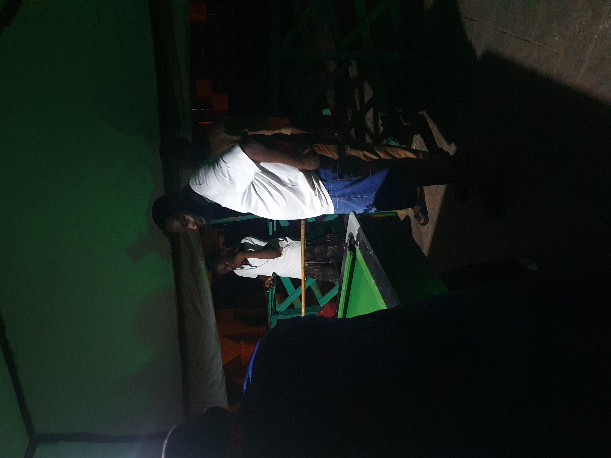 The image depicts an indoor scene, possibly a bar or recreational center, with a green-lit ceiling and dark surroundings. In the foreground, a person wearing a dark blue shirt is partially visible from the back, facing away from the camera. To the right, a man in a white t-shirt and blue jeans stands, looking towards a pool table. Another person in a light-colored t-shirt is positioned behind the pool table, seemingly playing or observing the game. The pool table itself has a green surface and is illuminated from above. The overall atmosphere is dim, with shadows cast by the people and the objects in the space. The image does not provide explicit details to determine its exact location within Lokoja, Nigeria, but it suggests a social gathering or entertainment setting.