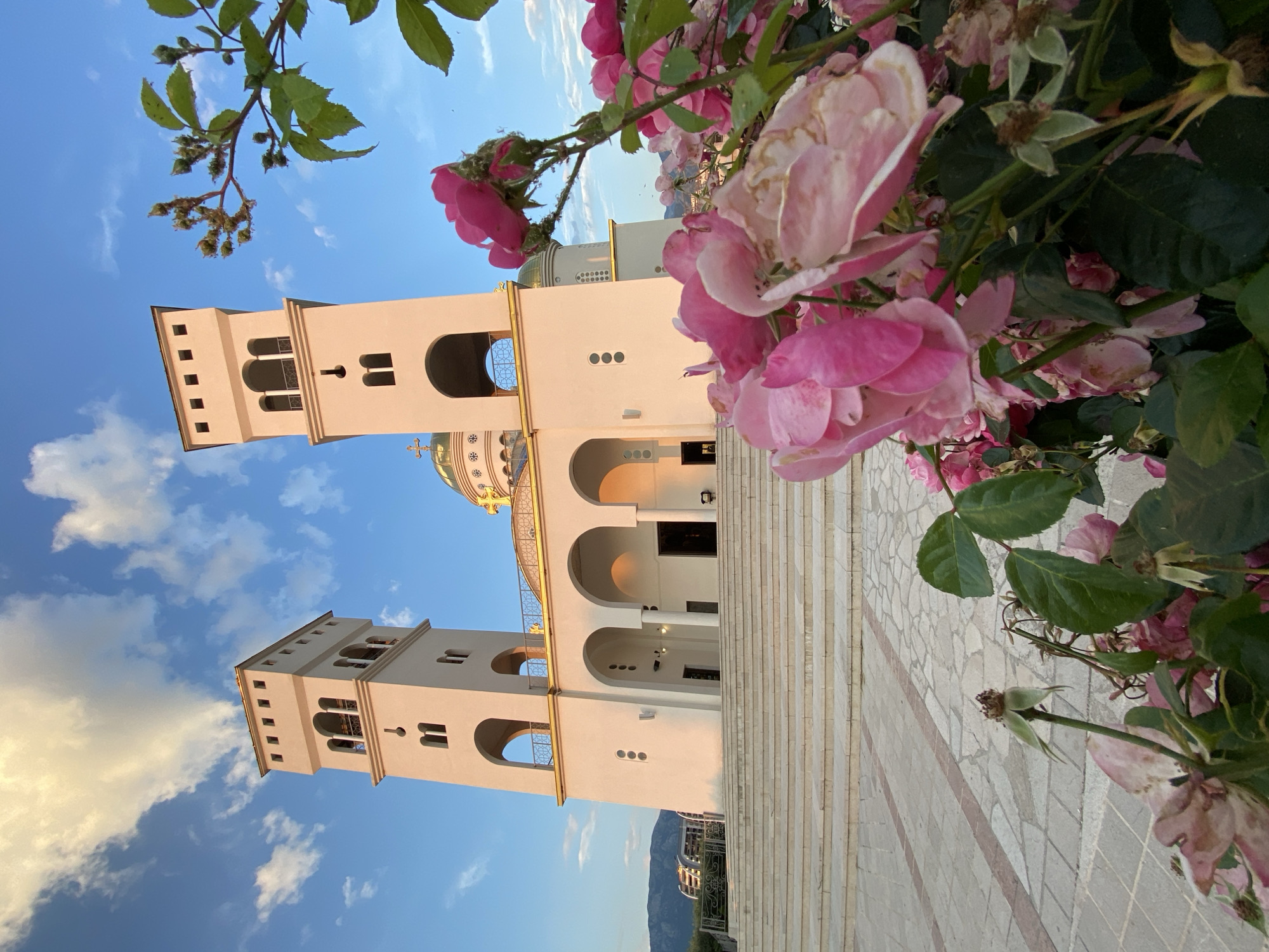 The image shows a low-angle, vertical shot of the Cathedral of Saint John Vladimir, located in Bar, Montenegro. The cathedral's white façade, featuring two prominent bell towers and arched entrances, is partially framed by pink roses in the foreground. The sky behind the cathedral is blue with scattered white clouds. The foreground also includes sections of a paved walkway leading up to the cathedral's entrance. In the background, there is a faint view of a mountain range. There are no people visible in the frame.