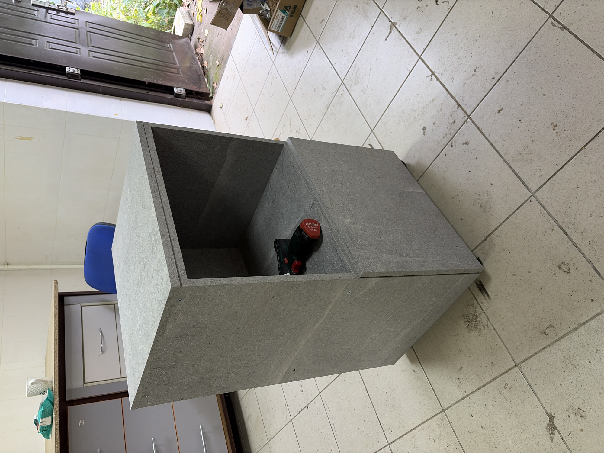 The image depicts an unfinished cabinet with a drawer and an open shelf, situated on a tiled floor. A black and red power drill is placed on the shelf. To the left of the cabinet is a filing cabinet with several drawers. In the background, a blue chair and a doorway leading outside are visible. The environment appears to be an indoor workshop or storage area. The scene is located in Nizhneye Khoroshovo, Russia.