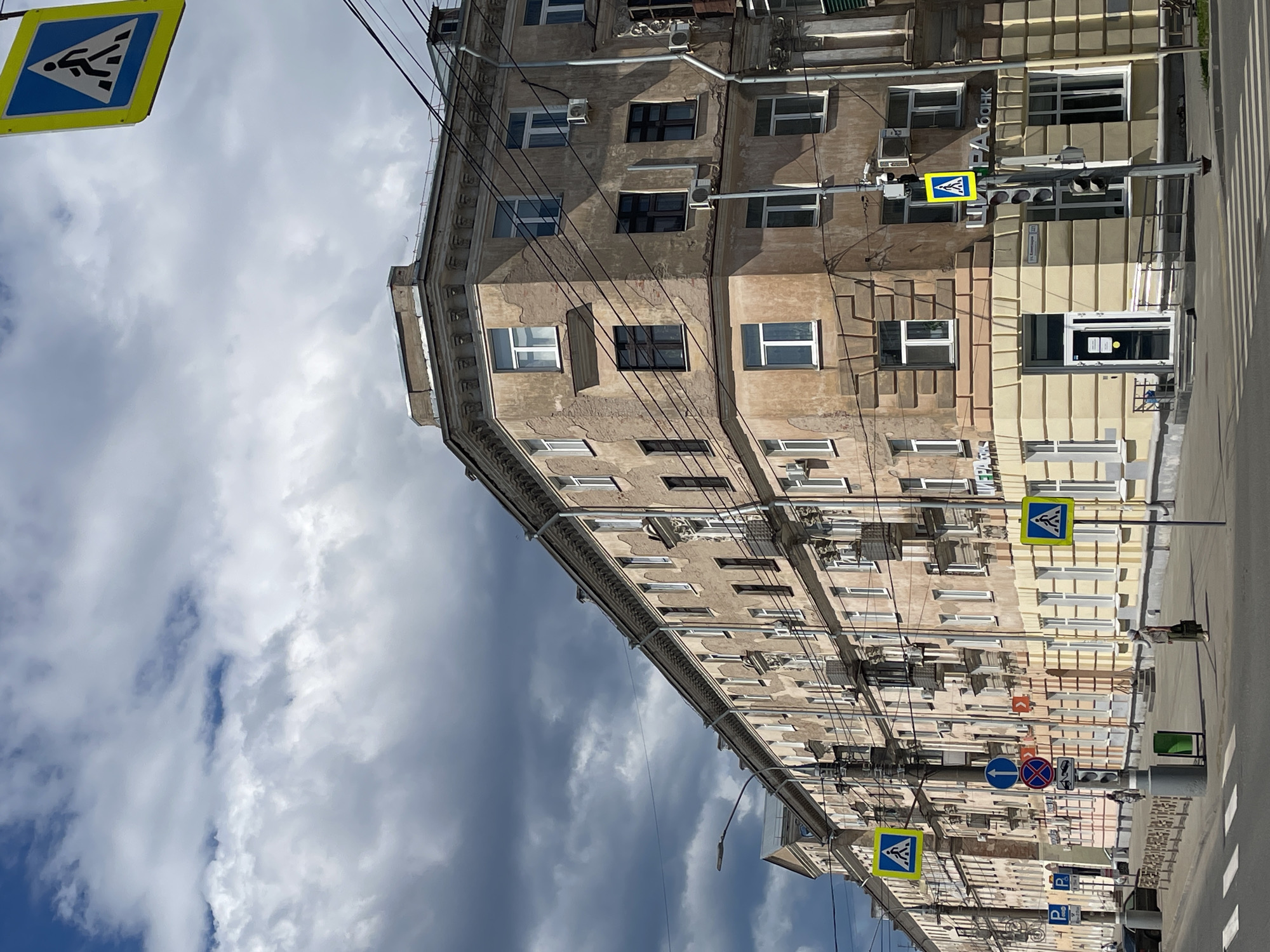 This is a photograph taken from a slightly elevated perspective, looking up at the side of an old, multi-story apartment building. The building is constructed with worn, light-colored brick and features numerous windows, many with white frames and some with dark, closed shutters. The architecture suggests an older European style, with decorative elements along the top of the building. Electrical wires are strung across the facade, adding to the urban feel.
The scene is set on a street with a visible pedestrian crossing. Several traffic signs are present, including a yellow diamond-shaped sign with a blue pedestrian silhouette, indicating a crosswalk. There are also traffic lights and other regulatory signs. The building's facade shows signs of age, with some discoloration and peeling paint. Air conditioning units are mounted on some of the walls.
The sky is partly cloudy, with a mix of bright white clouds and darker, more ominous clouds, suggesting a dynamic weather day. The lighting indicates it's daytime, possibly late morning or afternoon, given the shadows. The overall mood is somewhat utilitarian and urban, with no specific event or activity clearly depicted, beyond the everyday presence of traffic and pedestrian infrastructure. No people are clearly visible in the frame. The visible text on the building is minimal and partially obscured, but appears to be a sign with Russian lettering that may indicate a business or organization.