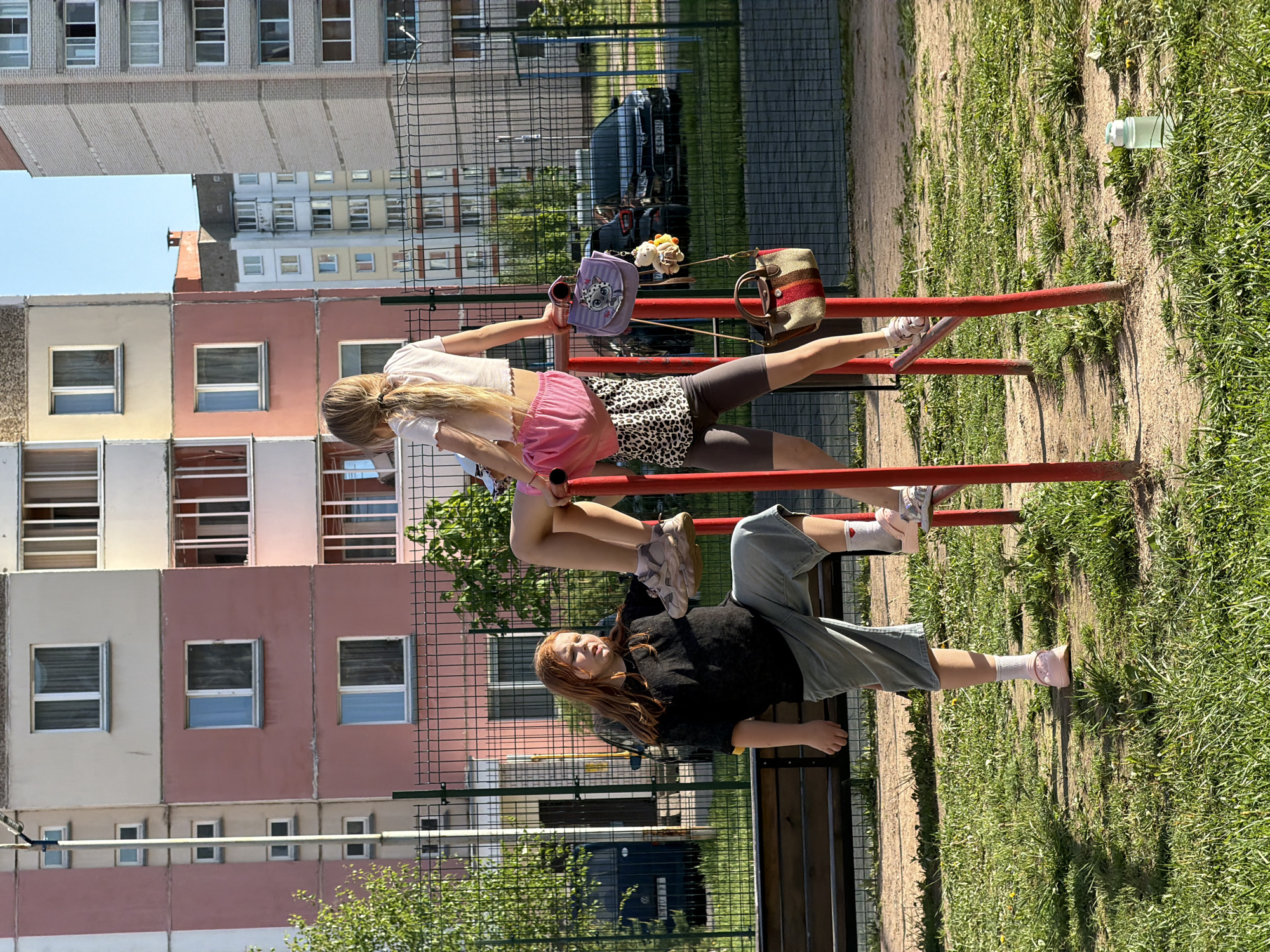 The image shows two girls on a red metal playground structure with a building in the background. One girl is standing on the structure, and the other is upside down with her feet up on it. Several bags are hanging from the structure. The playground is adjacent to a building. A metal fence separates the playground from the building.