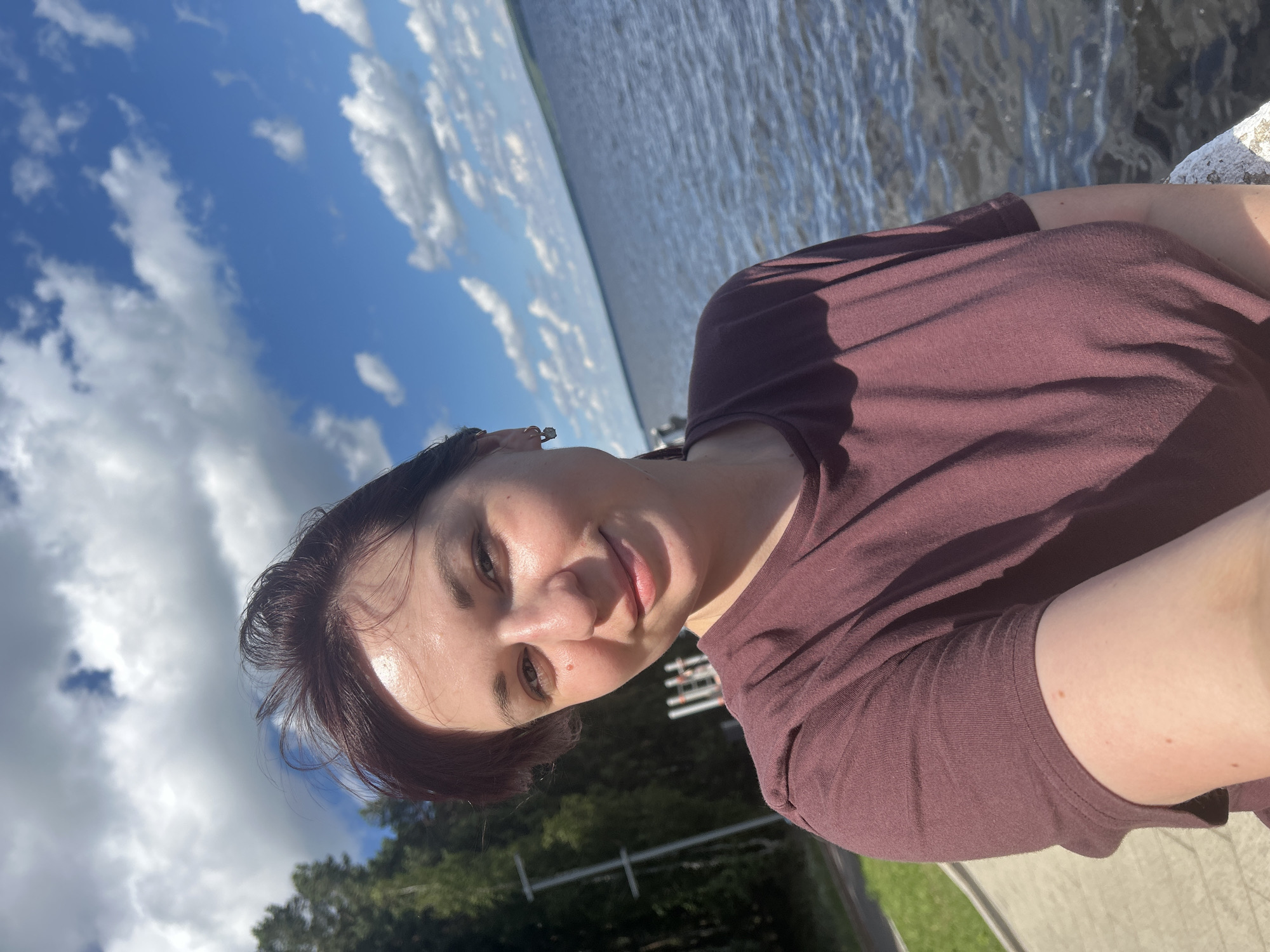 A portrait of a young woman is taken from a low angle, with the camera tilted. She has short, dark hair and is wearing a maroon t-shirt. The woman is smiling and looking towards the camera. Her skin is dewy and appears to be illuminated by sunlight. She is wearing a small earring in her left ear.
The background of the image consists of a body of water that reflects the bright blue sky and white clouds. Trees can be seen along the distant shore, suggesting a natural outdoor setting. The overall lighting and clear sky indicate it is daytime and the weather is fair. The image seems to be a selfie taken by the woman herself, possibly while outdoors near a lake or river. The location is suggested to be Chaikovsky, Russia.