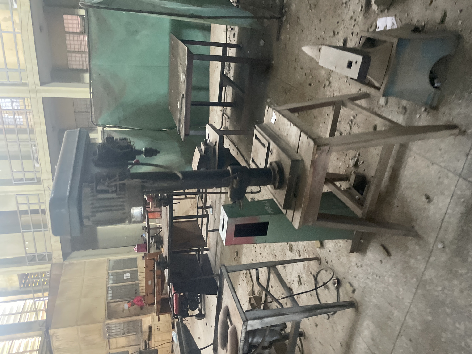 This image depicts a workshop environment in Ihiagwa, Nigeria, likely within a technical or vocational training facility. In the foreground, a large drill press stands prominently on a wooden stool. To its left, a metal workbench holds two elongated metal objects. Nearby, a green stand with a red-lined opening is positioned. An anvil on a blue base is situated to the right of the drill press. The workshop floor is concrete and littered with debris. In the background, several tables are visible, some covered with a green tarp. One table has tools on it, including what appears to be a small vise. Further back, a red generator is present, and a person wearing a red shirt is visible through a window. The overall environment suggests a functional but somewhat cluttered workspace.