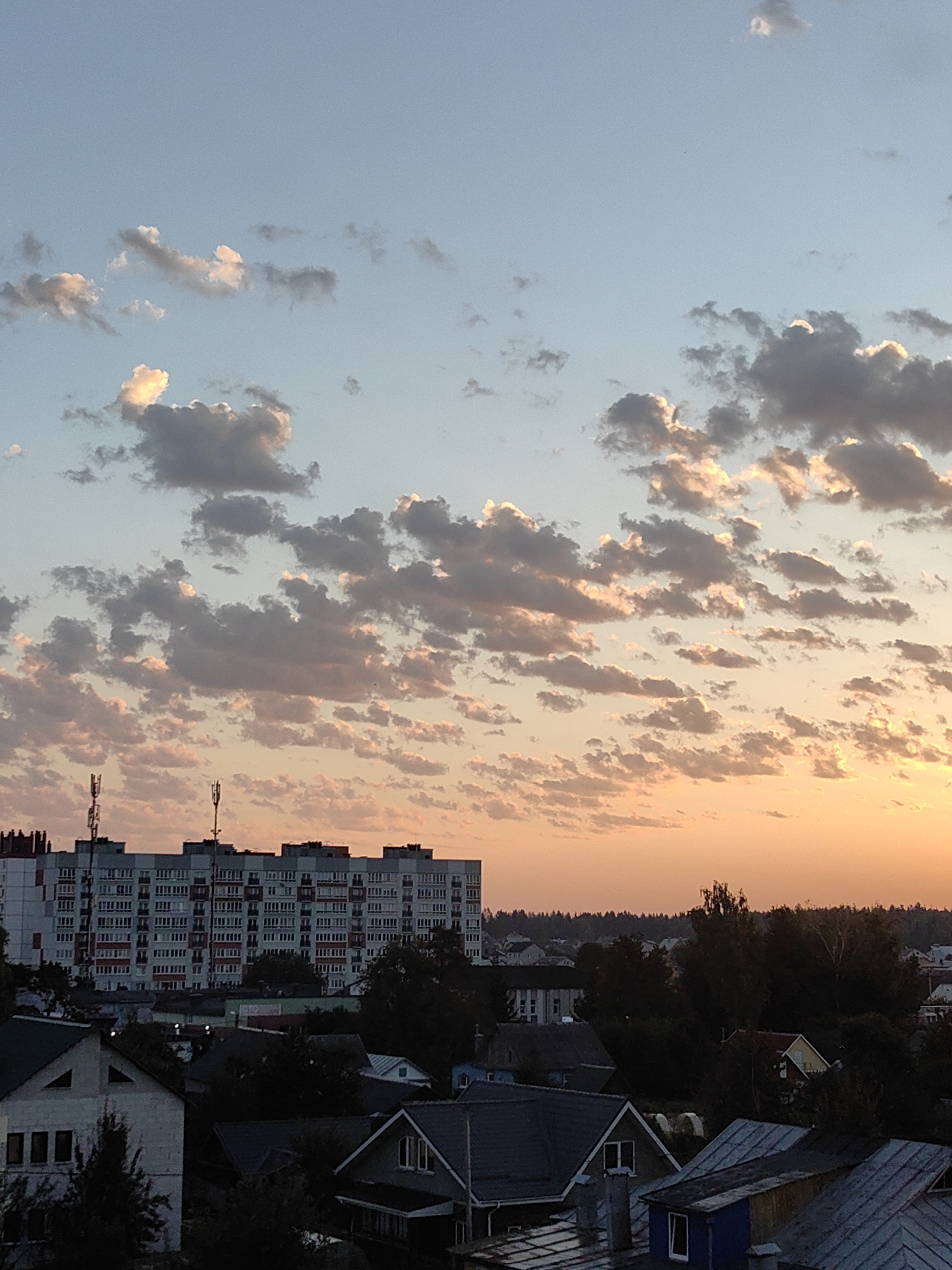 This image depicts a cityscape at sunset, likely in Fanipal, Belarus. The sky is filled with scattered, fluffy clouds, tinged with orange and pink hues from the setting sun, which is partially visible near the horizon. Dominating the mid-ground is a large, multi-story apartment building with numerous windows and balconies, and two telecommunication towers on its roof. In the foreground and lower part of the image, a collection of houses with pitched roofs and trees are visible, suggesting a residential area. The overall scene captures the transition from day to evening in a European town.