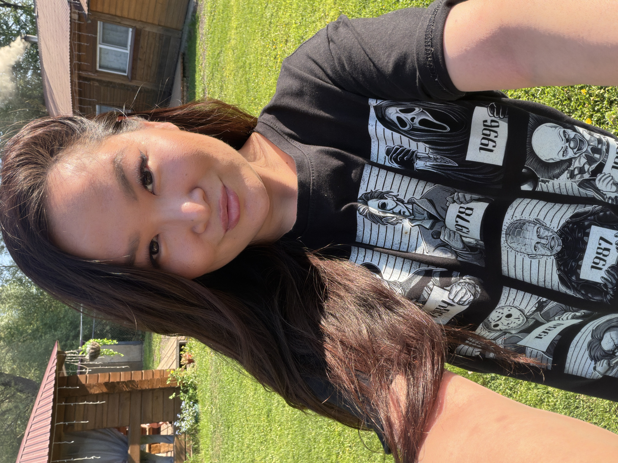 A woman is taking a selfie outdoors, likely in Malyi Bukor, Russia, given the prompt. She is wearing a black t-shirt with images of horror movie villains (Ghostface, Pennywise, Pinhead, Jason Voorhees, Freddy Krueger, and Michael Myers) and their associated release years. She is positioned on a grassy lawn, with a wooden structure with a brown roof visible in the background on the left side of the frame, and another similar building farther in the background on the upper left corner of the image. The lighting suggests it is daytime with sunny conditions.