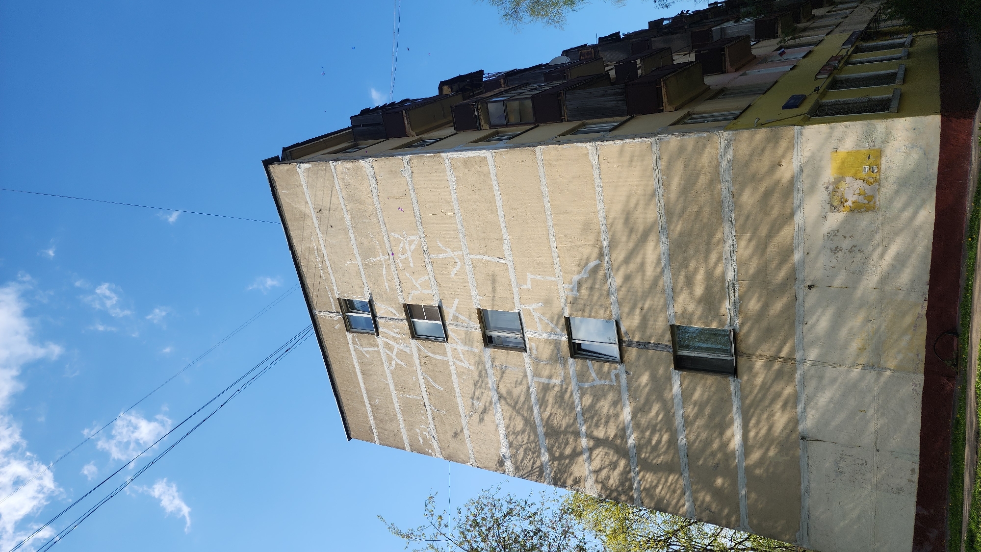 The image shows the exterior of a Soviet-era apartment building in Zelenograd, Russia, viewed from a low angle. The building is multi-story, constructed of prefabricated concrete panels, and painted in shades of tan and yellow. There are several rectangular windows on the visible facade, some with reflective glass. Vertical concrete seams divide the panels. The building shows signs of weathering and graffiti. The top floors have projecting balconies or enclosed loggias. Overhead power lines cross the clear blue sky in the background. Green trees are visible at the base of the building.