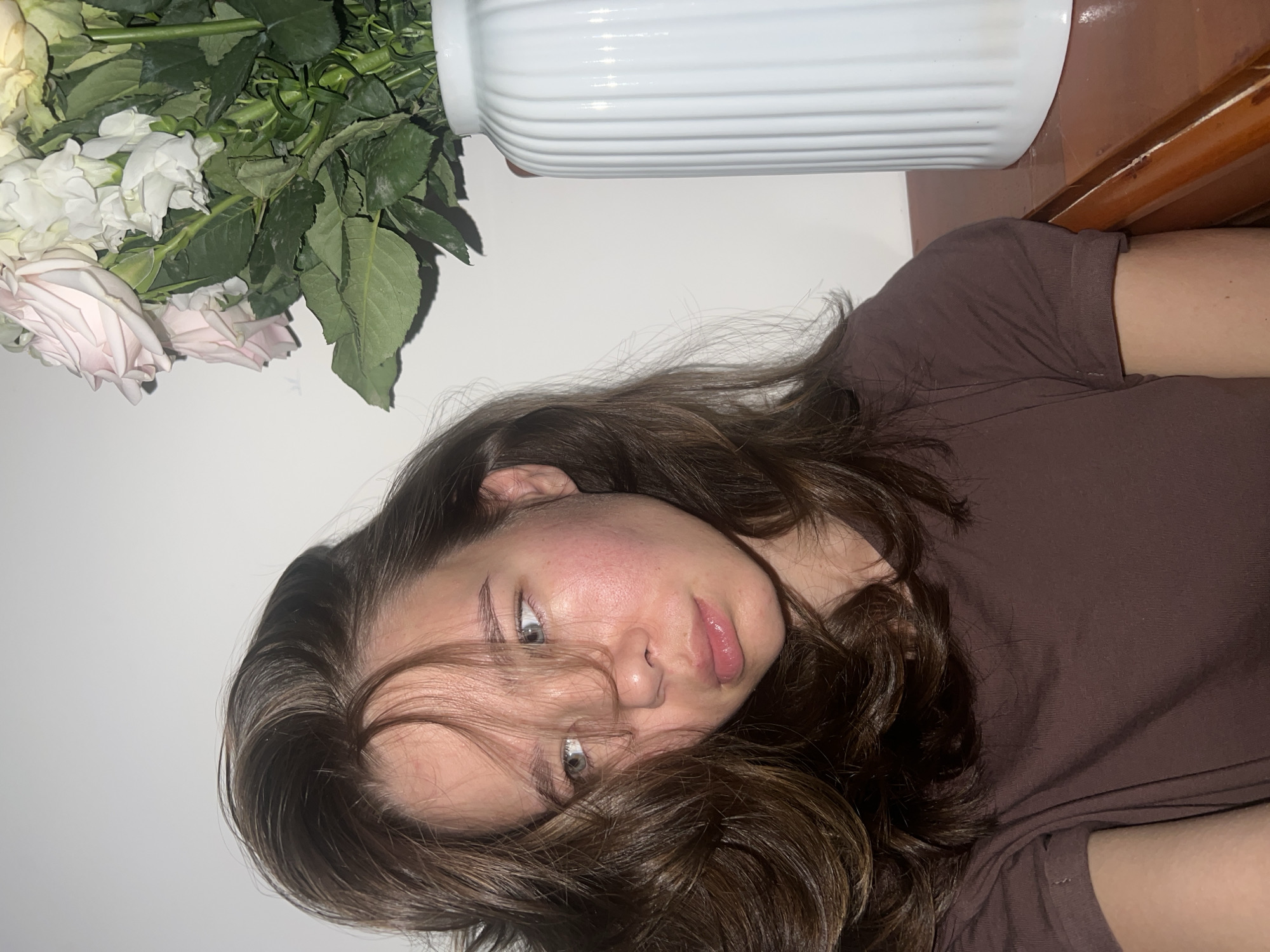 A woman with light skin, dark hair, and light eyes is lying down. Her head is tilted towards the viewer, and some of her hair falls across her face. She is wearing a brown t-shirt. Above and to the left of her head is a vase of flowers with white and light pink blooms and green leaves. The vase is white and ribbed. A portion of a brown wooden surface is visible to the right of the vase. The background is a plain white wall. The location is not evident from the image.