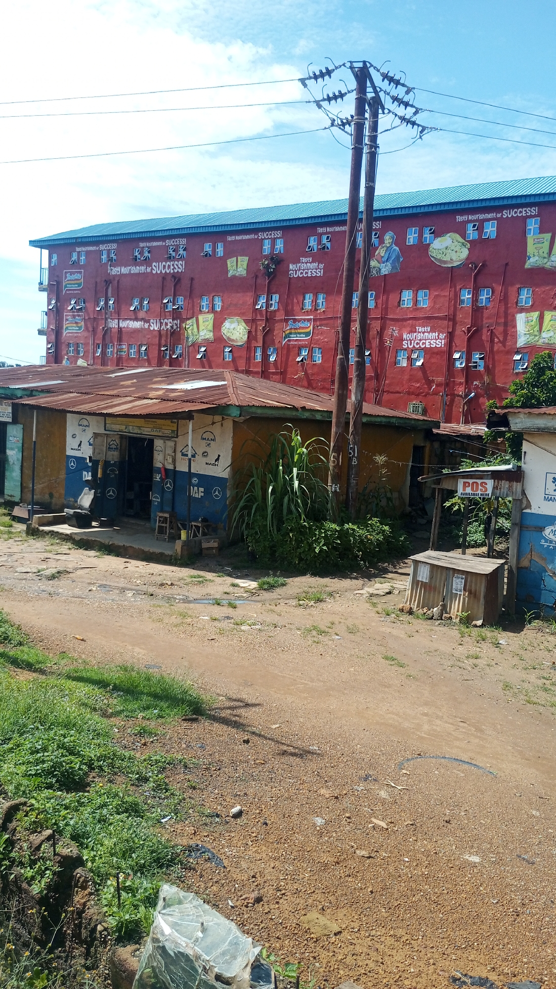 The image depicts an exterior scene in Rafin Par, Nigeria. In the background, a large red building with a blue roof is visible, adorned with numerous advertisements for "Tasty Nourishment for SUCCESS!" and pictures of food products. A prominent utility pole with electrical wires stands in front of the building. In the foreground, there are several small structures, including a shop with a rusty tin roof displaying logos for "MAN" and "DAF," and a smaller stall with a sign that reads "POS AVAILABLE HERE." The ground is covered in dirt and gravel, with some patches of green grass and vegetation. The overall impression is of a developing urban or semi-urban environment. No people are visible in the scene.