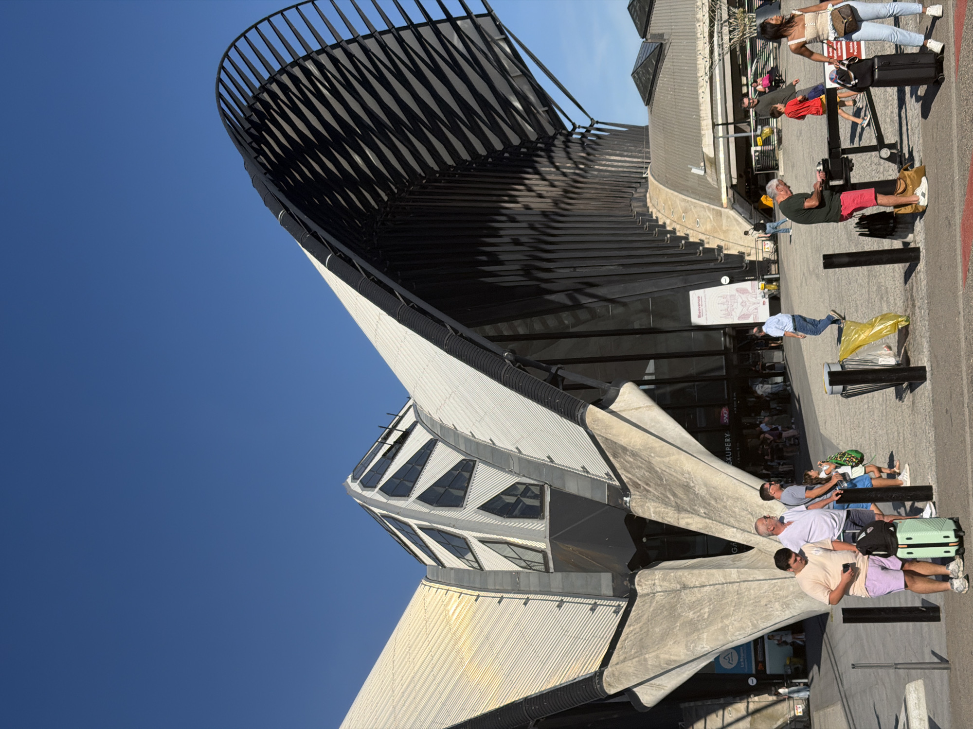 The image captures the exterior of Lyon-Saint Exupéry Airport, located in Colombier-Saugnieu, France, under a clear blue sky. The distinctive architecture features a large, curved, dark-slatted roof extending over a white, ribbed structure with triangular windows. In the foreground, several individuals are present, some with luggage. A young man in a light-colored t-shirt and lavender shorts stands looking at his phone, with a mint-green suitcase and a black backpack at his feet. Beside him, an older man in a white t-shirt and blue shorts stands. Further to the right, a man in a green t-shirt and red shorts is engaged with a table or stand. A woman in a white top and blue jeans stands with a black suitcase. Other people are visible in the background, some entering or exiting the building, which displays signage including "Bienvenue" and "SNCF". The ground surface is paved, and there are black bollards present.