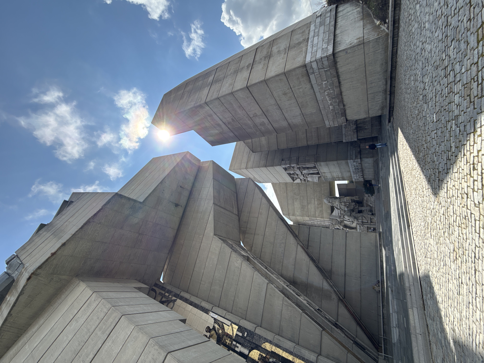 The image shows the Founders of the Bulgarian State Monument in Shumen, Bulgaria. The monument features several monumental concrete structures with sharp angles pointing towards the sky. The sun is shining, partially obscured by clouds, with a bright glare visible between two of the structures. A cobblestone surface is visible to the side of the monument. The monument features a number of sculptures. There are a few people visible near the base of the monument. The sky is blue with scattered white clouds.