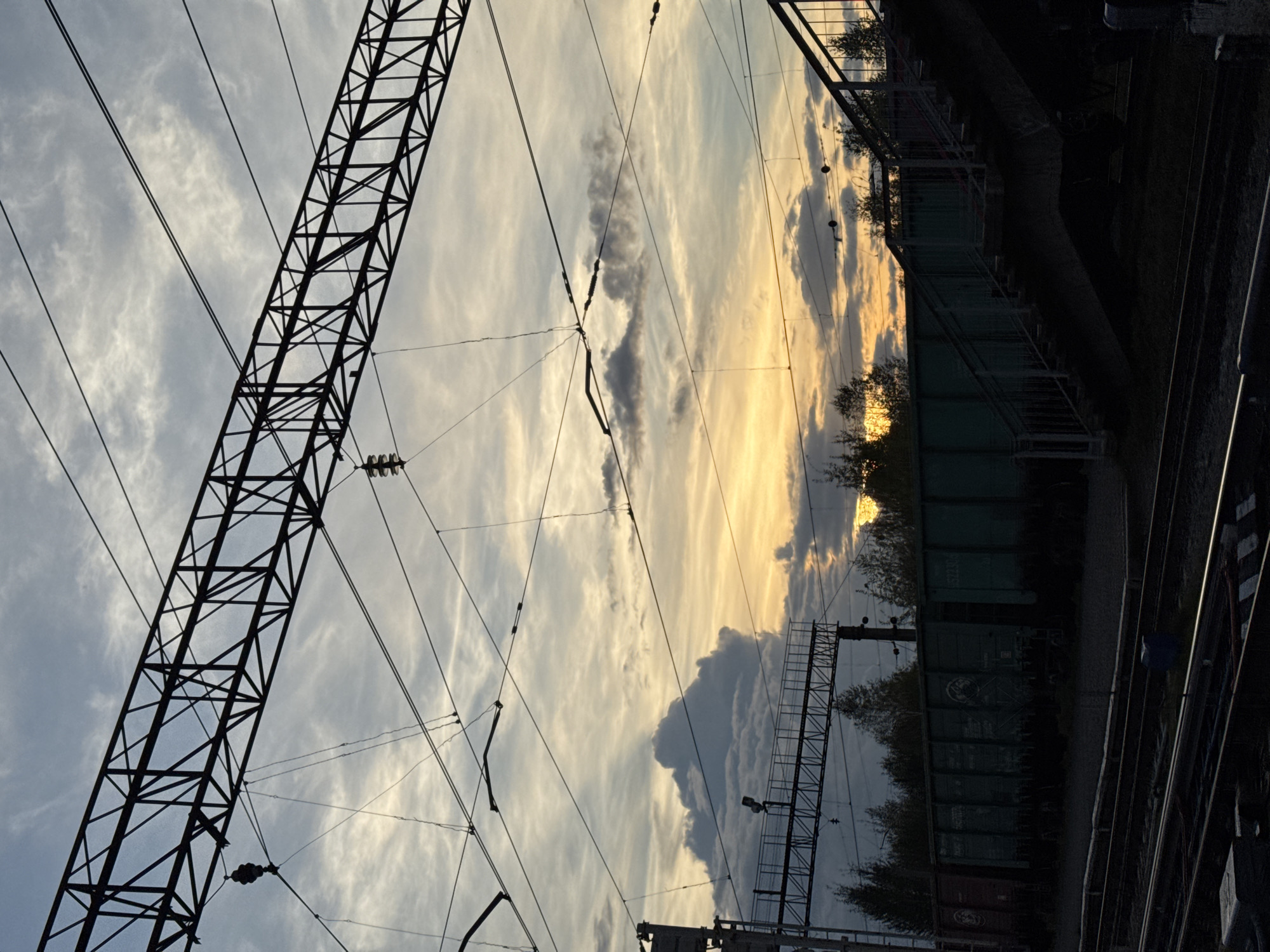 The image depicts a railway yard at dusk. The sky is filled with dramatic clouds, illuminated by the setting sun, casting warm yellow and orange hues. Overhead, a complex network of power lines and supporting structures crisscrosses the frame, silhouetted against the sky. In the middle ground, a row of green freight cars is parked on the tracks. To the right, a pedestrian overpass with stairs provides access to a platform. The foreground shows multiple sets of railway tracks. The overall impression is one of industrial activity against a natural, atmospheric backdrop. The scene is set in Komarikha, Russia.