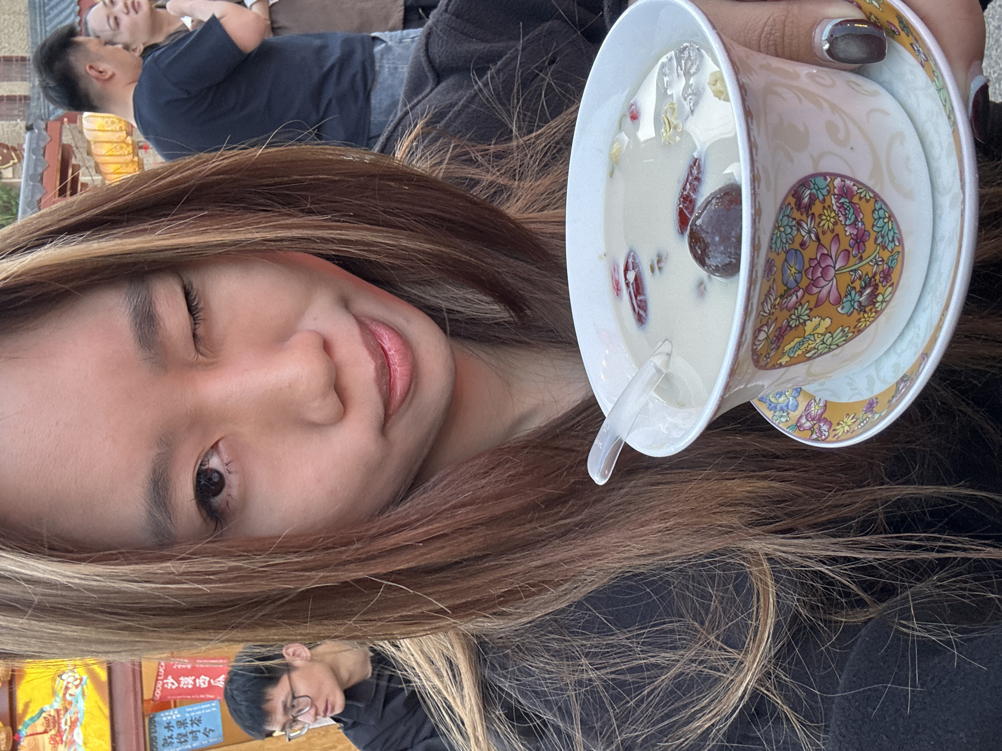 A woman with shoulder-length highlighted brown hair is centered in the image, holding a bowl of what appears to be dessert in her right hand. She is wearing a black shirt and is winking her left eye at the camera. The bowl has a white interior, and the dessert consists of a milky liquid with what looks like red dates and other ingredients. The exterior of the bowl has a floral pattern in shades of yellow, orange, and pink.
In the upper left corner of the frame, two men are visible. One is wearing a navy shirt and is partially out of focus. The other is leaning on him, and only a portion of his head is visible. In the lower left corner, another man is partially visible, wearing glasses and a black shirt. There is a sign in Chinese characters partially visible to the left of this man.
The overall impression is a casual selfie taken in a public place. The presence of the Chinese sign and the dessert being consumed suggest a location in China. Given the specification of Dunhuang City, China, it can be inferred the scene is likely within that city, possibly at a restaurant or outdoor dining area.