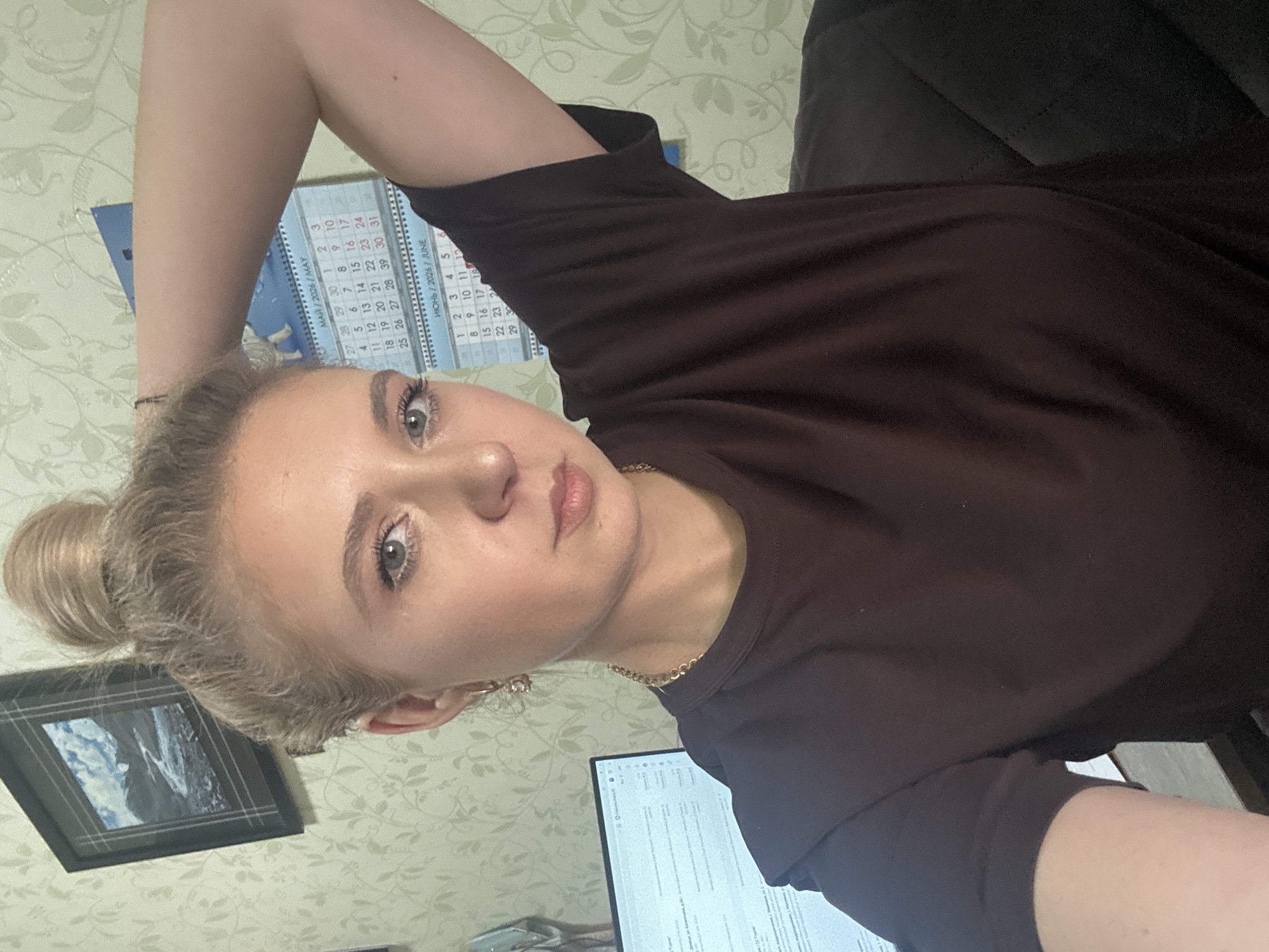 A young woman with blonde hair pulled up in a bun is positioned in a close-up, waist-up shot. She is looking at the camera with a neutral expression, her left arm raised behind her head and her right arm partially visible. She is wearing a dark brown t-shirt and a gold chain necklace. In the background, a calendar with the dates "March/2026/May" and "June/2026/June" is visible on a wall with a floral wallpaper pattern. To the left of the woman, a computer monitor is partially visible, displaying text. A framed picture of a landscape is hanging on the wall above and to the left of the woman. The scene appears to be an indoor setting, possibly an office or home environment, as indicated by the computer and personal items.