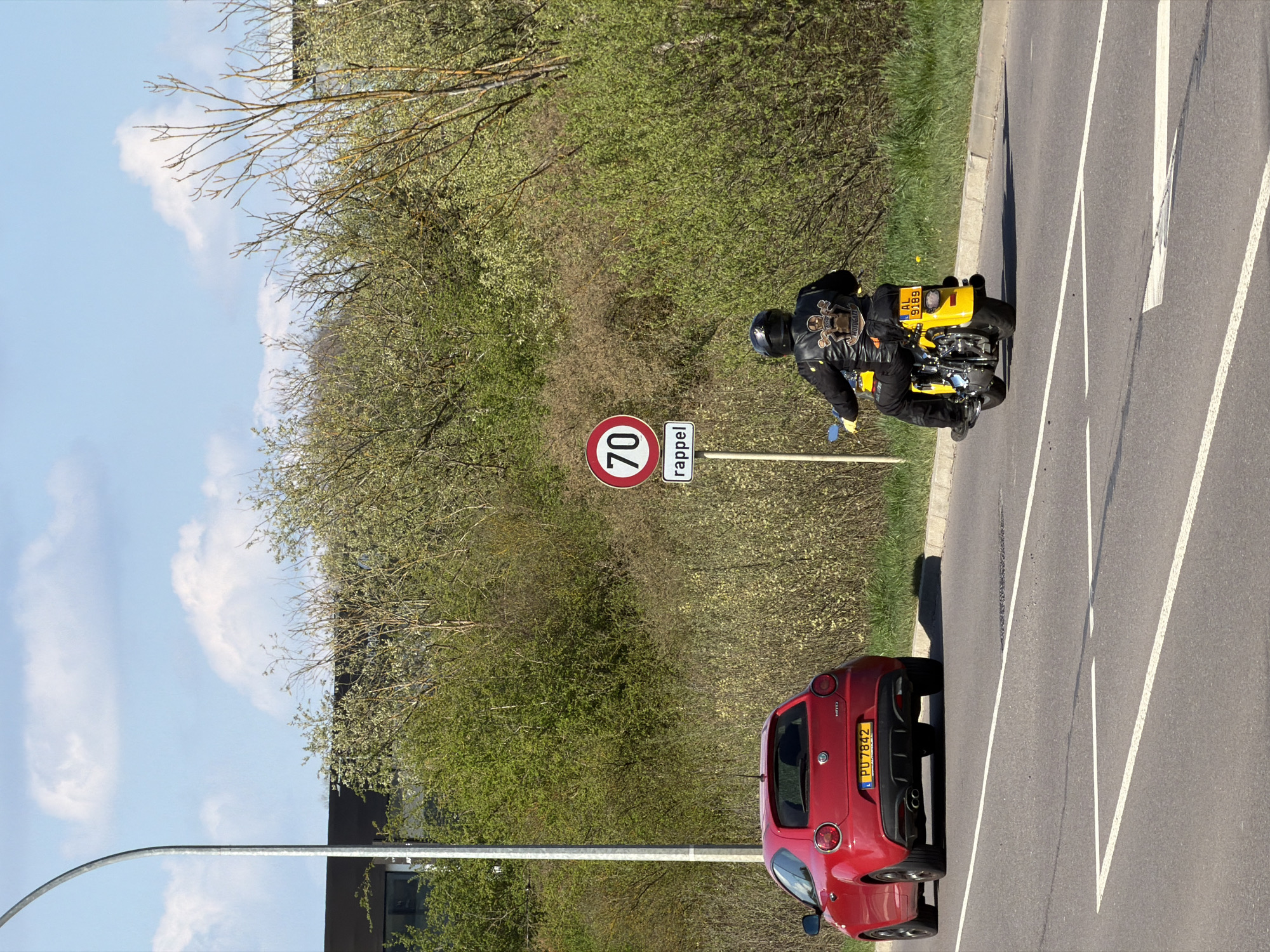 The image shows a street scene in Esch-sur-Alzette, Luxembourg. A motorcyclist wearing a helmet and black leather jacket rides a yellow motorcycle with a Luxembourg license plate reading "AL 5969". To the left of the motorcyclist, a circular red and white speed limit sign displays "70" with "rappel" (reminder) below. A red Alfa Romeo Mito with a license plate "PU 7842" is parked on the side of the road. The street has white lane markings and is flanked by green foliage on one side. In the background, there are trees, a building, and a blue sky with white clouds. A tall, metal street light pole is visible in the lower left corner of the frame.