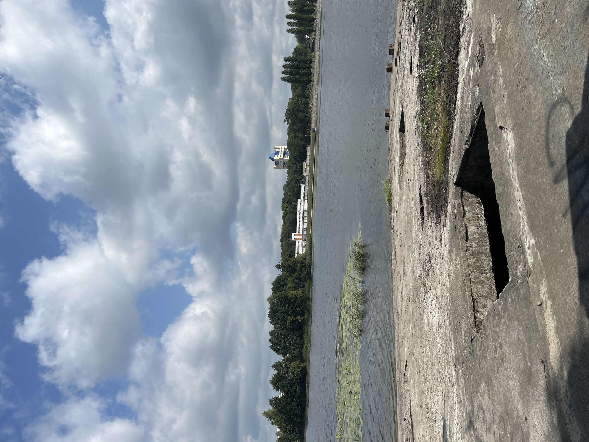 This image depicts a waterfront scene in Brest, Belarus. In the foreground, a cracked and weathered concrete surface with several square openings dominates the frame. Beyond this surface lies a calm, dark body of water, partially covered by patches of green aquatic vegetation. The middle ground features a densely forested treeline on the opposite bank of the water, interspersed with buildings. Notably, a modern building with a blue, pyramidal roof structure stands out against the greenery. The background is filled with a sky filled with dramatic, fluffy white and grey clouds against a bright blue sky. The overall impression is a somewhat industrial or abandoned waterfront area with a natural element of trees and water.
