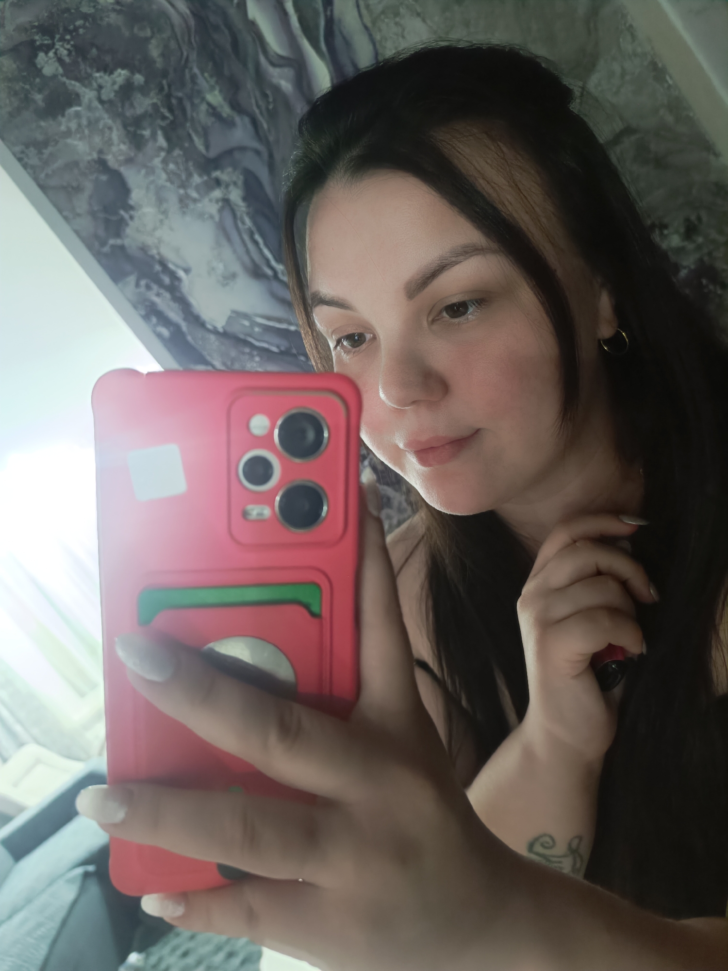 A woman is holding a red smartphone with a green cardholder on the back and taking a selfie. She has long, dark hair, a gold hoop earring in her left ear, and is wearing what appears to be a black top. Her nails are painted white. There's a glimpse of a tattoo on her left forearm. The background includes a marble-patterned wall or surface, a window with sunlight streaming in, and a portion of a gray couch. The image location is Chaikovsky, Russia.
