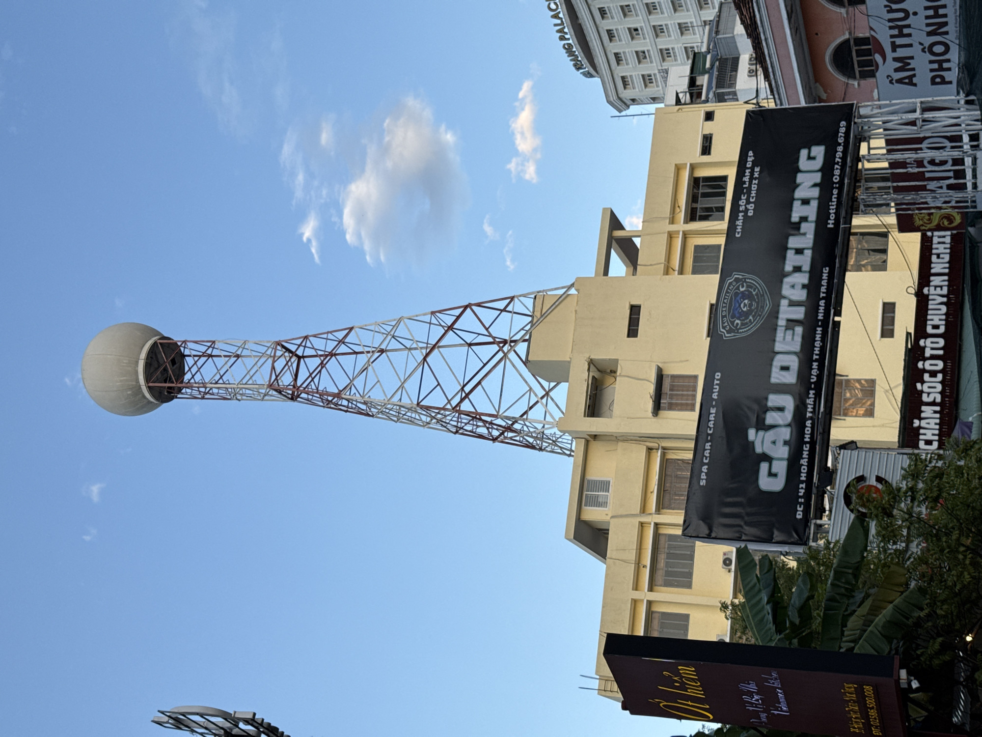 The image shows a tall radio tower with a spherical structure at the top, situated in front of a yellow building. Several signs are visible, including one that reads "GAU DETAILING" and another with the text "NHA TRANG" indicating the location. The building appears to be a commercial establishment, possibly with multiple floors. The sky is blue with a few scattered clouds, suggesting daytime. The overall scene is urban, with a mix of modern and older architecture.