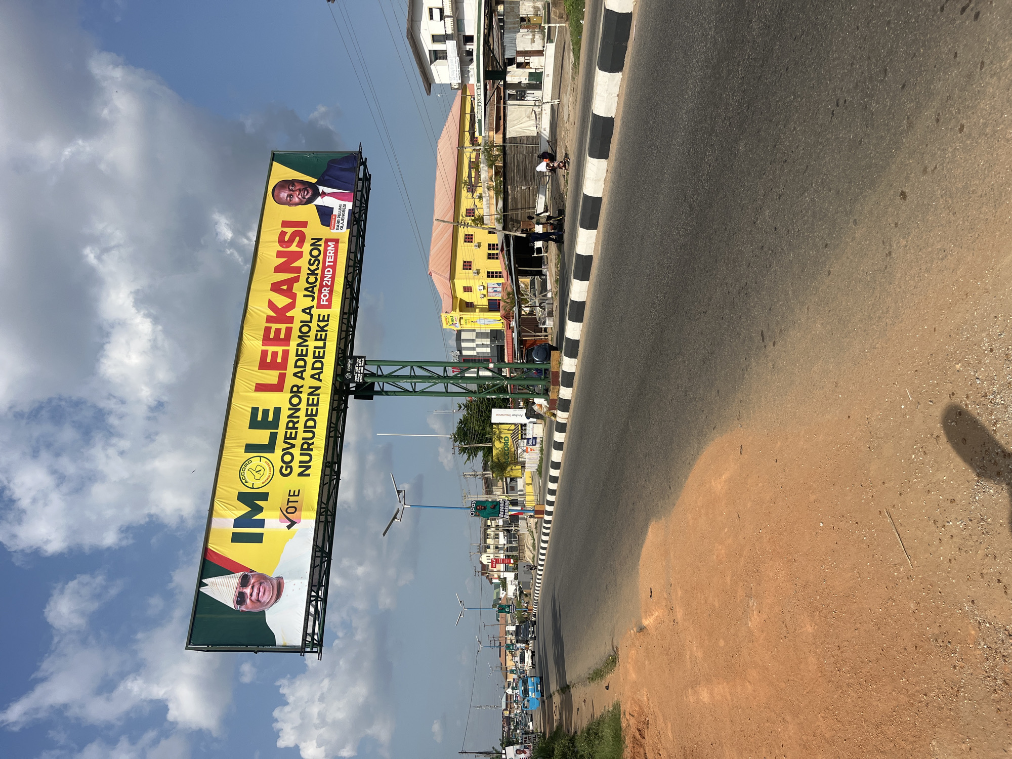 The image shows an outdoor scene in what appears to be Abeere, Nigeria. The main subject is a large billboard promoting the candidacy of "IMOLE LEEKANSI," labeled as "GOVERNOR ADEMOLA JACKSON NURUDEEN ADELEKE FOR 2ND TERM," alongside BARR PELUMI OLAJENGBESI. The billboard is situated beside a road with black and white painted curbs. In the background, there are buildings, including a yellow structure. There is a glimpse of a person standing by the side of the yellow structure with another person by their side and vehicles and street lighting further down the road. The sky is visible with clouds. The image is oriented at an angle, with the road running from the top right corner towards the bottom left.