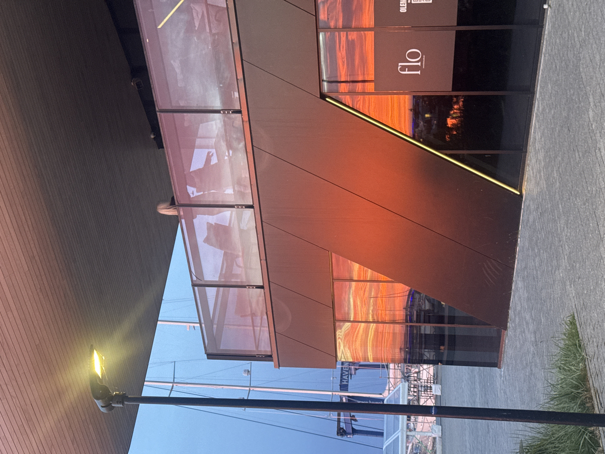 The image shows a modern building exterior at a harbor, likely in Tallinn, Estonia, during sunset. The building has a dark, angled facade with large glass windows that reflect the vibrant orange and yellow hues of the sky. A sign on one of the windows reads "flo." Above the dark facade, a glass-enclosed upper level is visible, with a few silhouettes of people and furniture suggesting it's a restaurant or bar area. A street lamp with a warm yellow glow is positioned to the left of the building, casting light downwards. In the background, the masts of sailboats and a crane indicate a port or marina setting. The sky is a gradient of deep blue and fiery orange, characteristic of a sunset. The ground in front of the building is paved with light gray cobblestones and bordered by a strip of green grass.