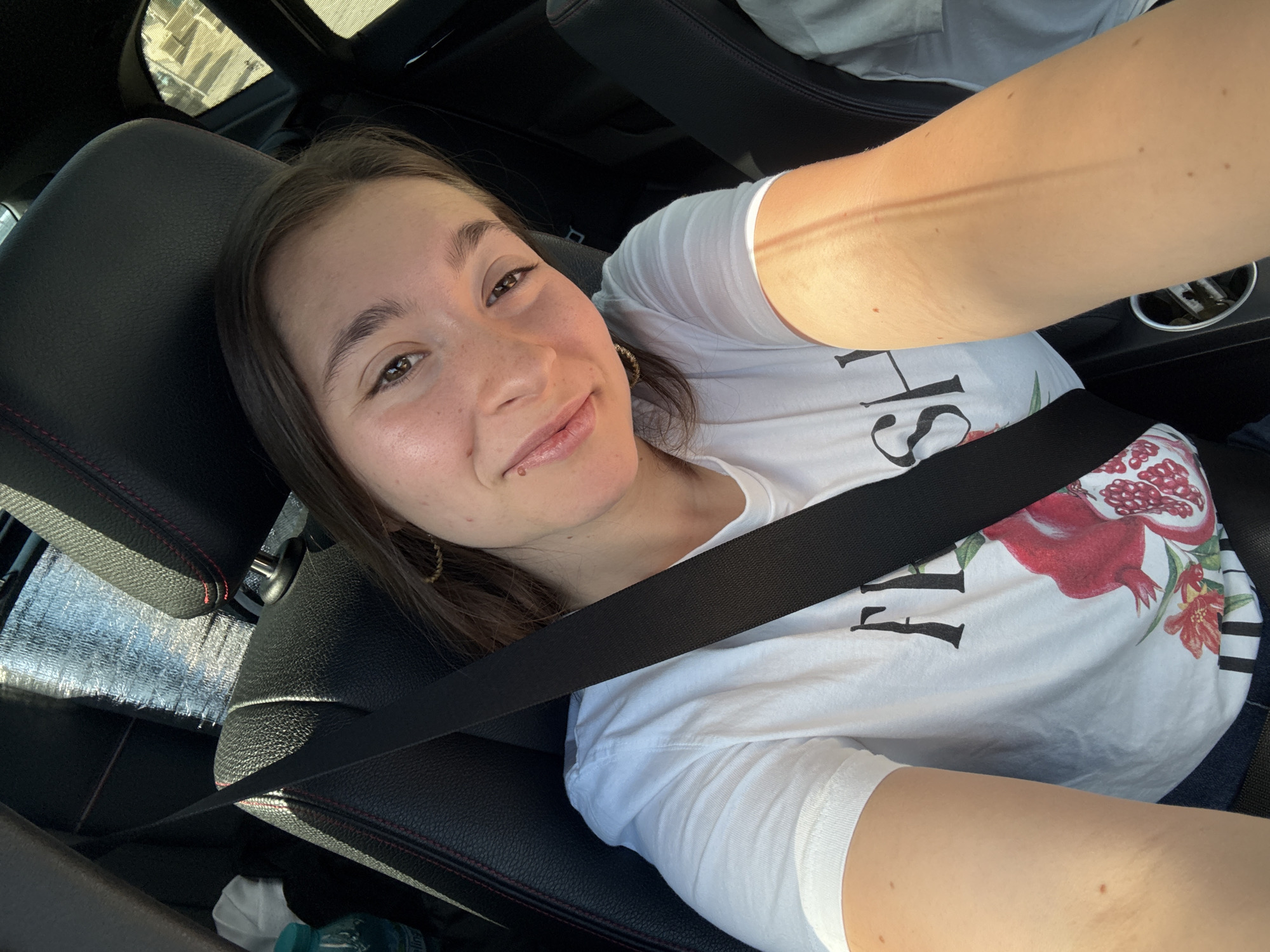 A young woman is taking a selfie from the passenger seat of a car, which is likely parked in Sofia, Bulgaria, given the context. She is wearing a white t-shirt with a graphic print of pomegranates and flowers, and a black seatbelt is fastened across her chest. The sun is shining brightly, casting a warm glow on her arm and face, suggesting it is daytime. The interior of the car is visible, with dark leather seats and red stitching, and a sunshade is positioned on the dashboard. The woman has a relaxed and happy expression, with a slight smile. Her hair is dark brown and appears to be shoulder-length. She is wearing a hoop earring in her visible ear. The overall mood is casual and pleasant.