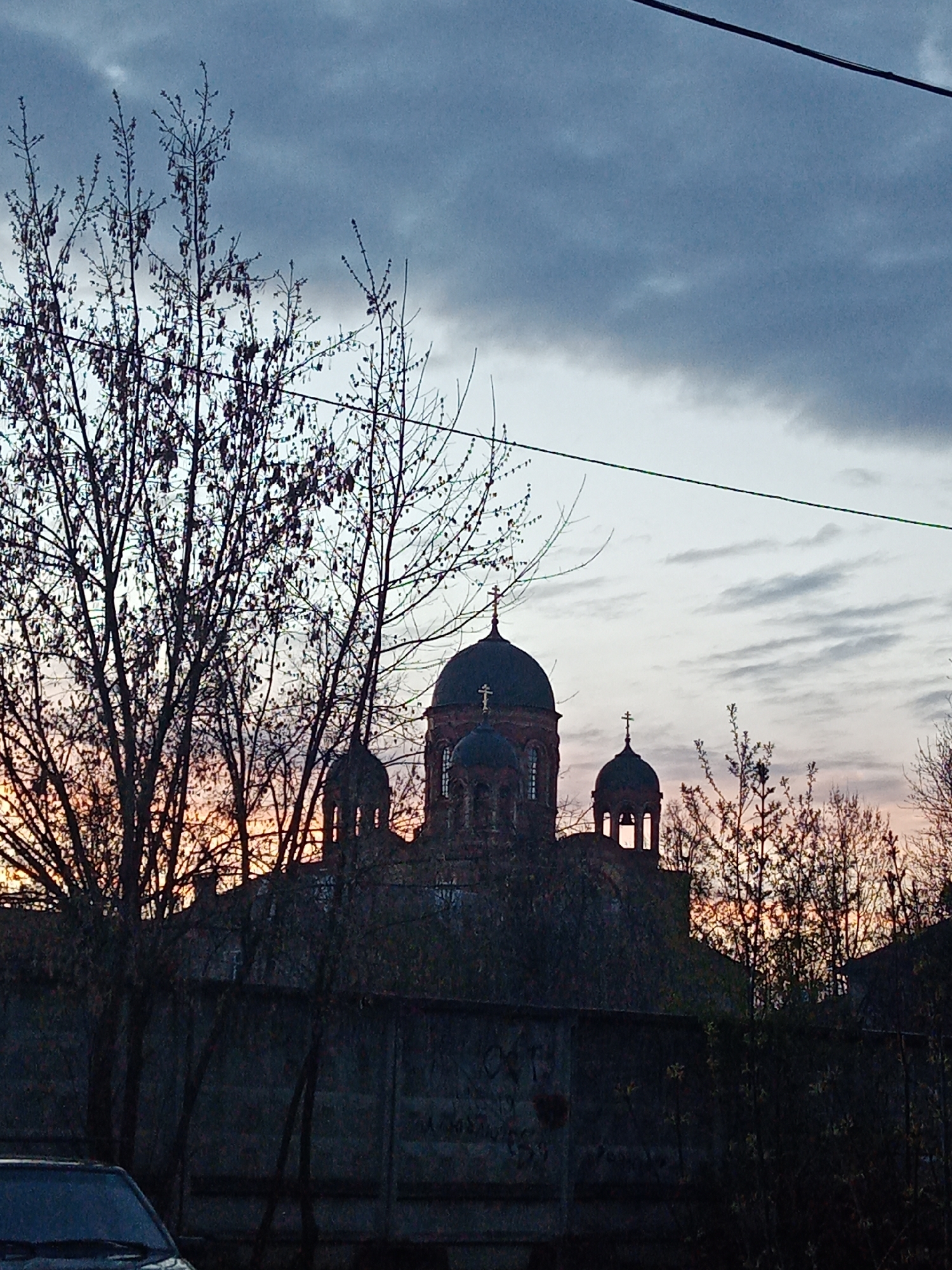 The image shows a building, identified as a church by the presence of crosses on its domes, located in Elektrostal, Russia. The church is silhouetted against a sky with varying cloud cover, indicating either sunrise or sunset. In the foreground, there are bare trees, partially obscuring the view of the church. A concrete wall or fence stands between the viewer and the church. A partial view of a vehicle is visible in the lower left corner. An overhead wire extends across the top of the frame.