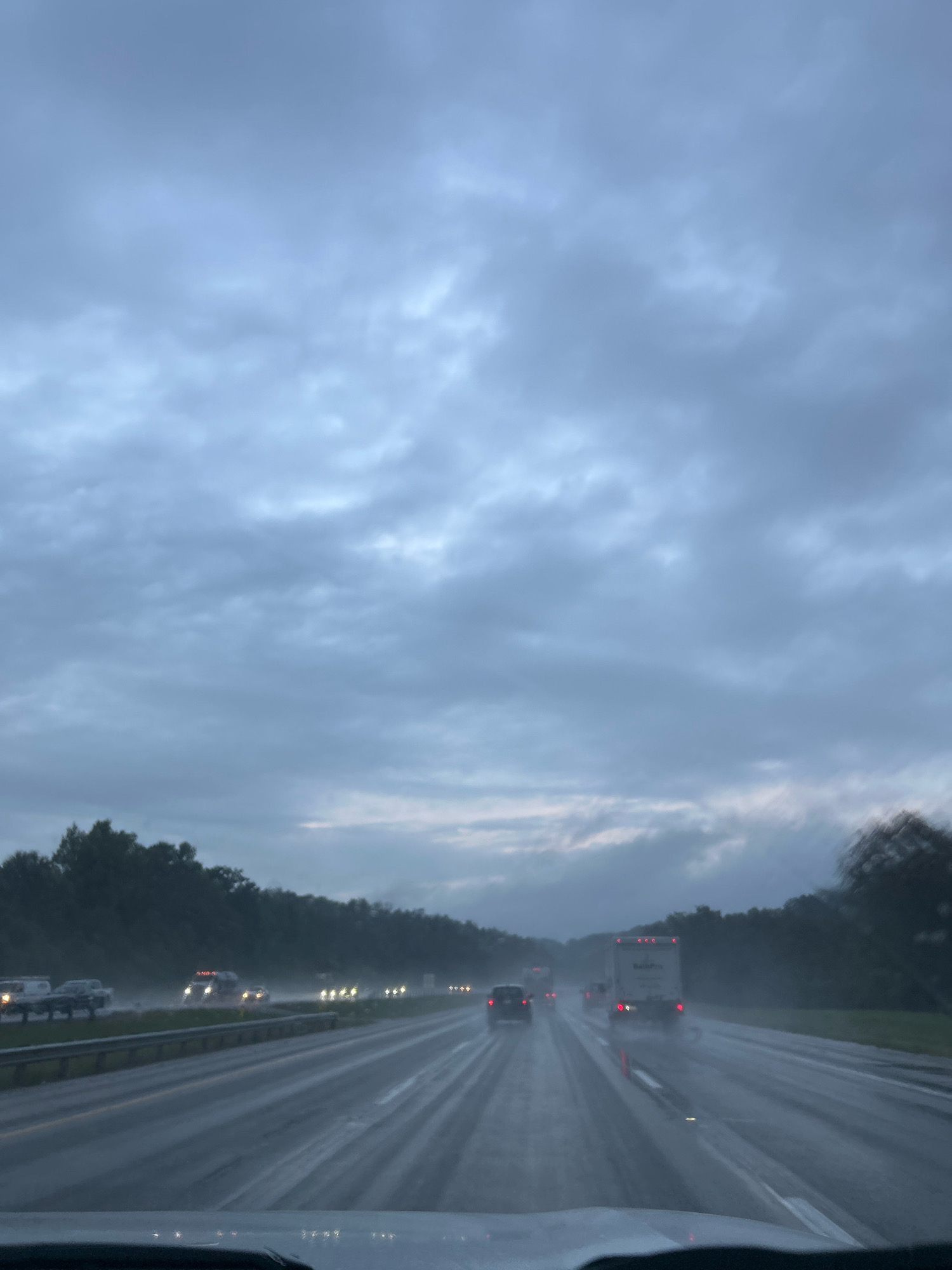 The image shows a view from inside a vehicle driving on a highway in Collier Township, United States of America, under a cloudy, overcast sky. The road is wet, indicating rain, and several vehicles, including cars and trucks, are visible on the highway, some with their headlights on. The surrounding environment consists of trees lining the sides of the highway. The overall scene depicts a rainy day drive on an American highway.