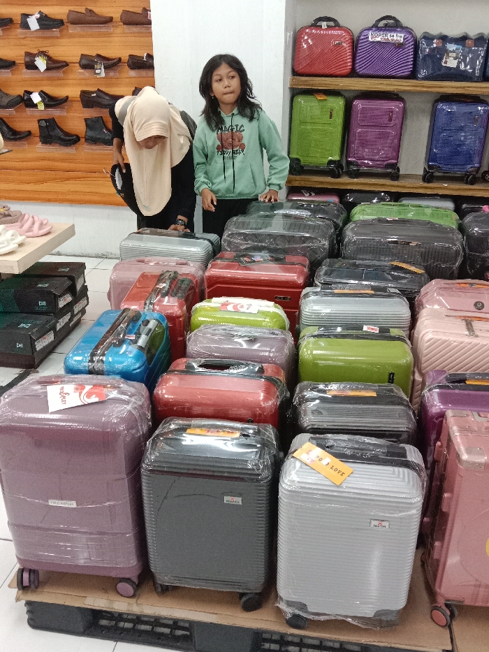 This image depicts an interior retail store in Langenharjo, Indonesia, specializing in luggage and footwear. In the midground, a young girl, approximately 8-12 years old, with dark, shoulder-length hair and wearing a light green hooded sweatshirt with a bear graphic, stands with her hands resting on a display of suitcases, looking towards the right. To her immediate left, an adult female, wearing a light-colored hijab and dark clothing, is partially visible, bent forward and examining items on a lower display.
The foreground and midground are filled with a dense arrangement of hard-shell suitcases of various sizes, primarily small to medium rolling cases and smaller cosmetic cases. These suitcases, predominantly wrapped in clear protective plastic, are displayed in multiple rows on low platforms or pallets. Visible colors include purple, grey, silver, red, blue, light green, and pink.
In the left background, a wooden display wall features numerous pairs of men's shoes, including loafers, boots, and dress shoes, arranged on multiple shelves. Below this shoe display, on a light-colored tabletop, are several pairs of light pink sandals or slippers. The right background contains additional displays of colorful suitcases on white shelves. The store has white walls and a light-colored tiled floor.