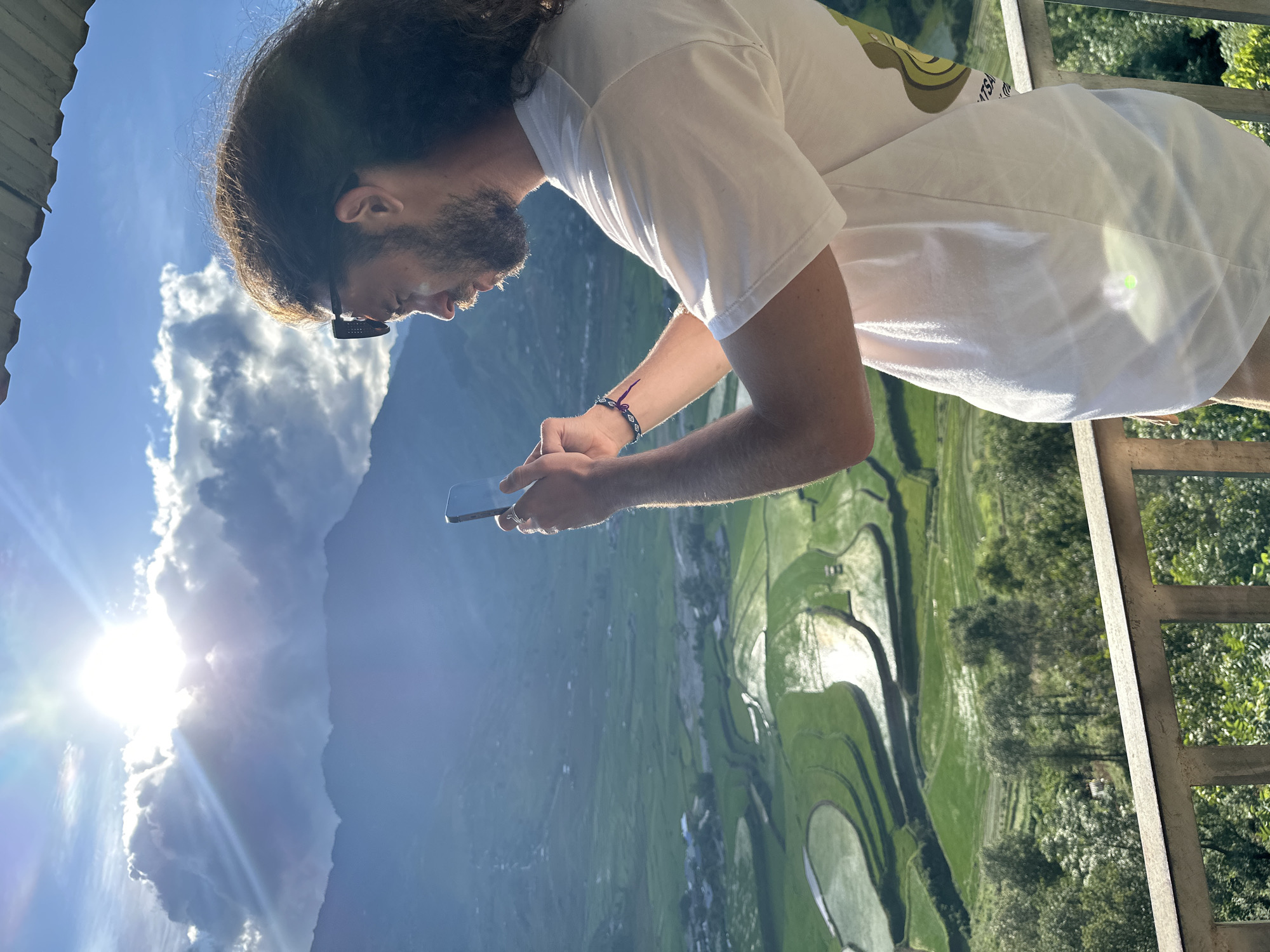 A man with long, dark curly hair and a beard, wearing a white t-shirt and sunglasses perched on his head, stands on a balcony in Tú Lệ, Vietnam. He is viewed in profile, facing right, and is holding a dark-colored smartphone with both hands, appearing to photograph the panoramic landscape. A blue and purple woven bracelet with a white eye-like bead is visible on his left wrist. The foreground features a light-colored balcony railing and the edge of a dark roof overhead. The background reveals a vast mountainous valley filled with vibrant green terraced rice fields, many reflecting the bright sunlight from their water-filled sections. Distant mountains rise under a bright blue sky with scattered clouds. The sun is intensely bright, positioned high in the sky to the left, casting strong light and creating lens flares. Small clusters of buildings are visible within the valley amidst the terraced fields. Lush green trees cover the slopes below the cultivated areas.