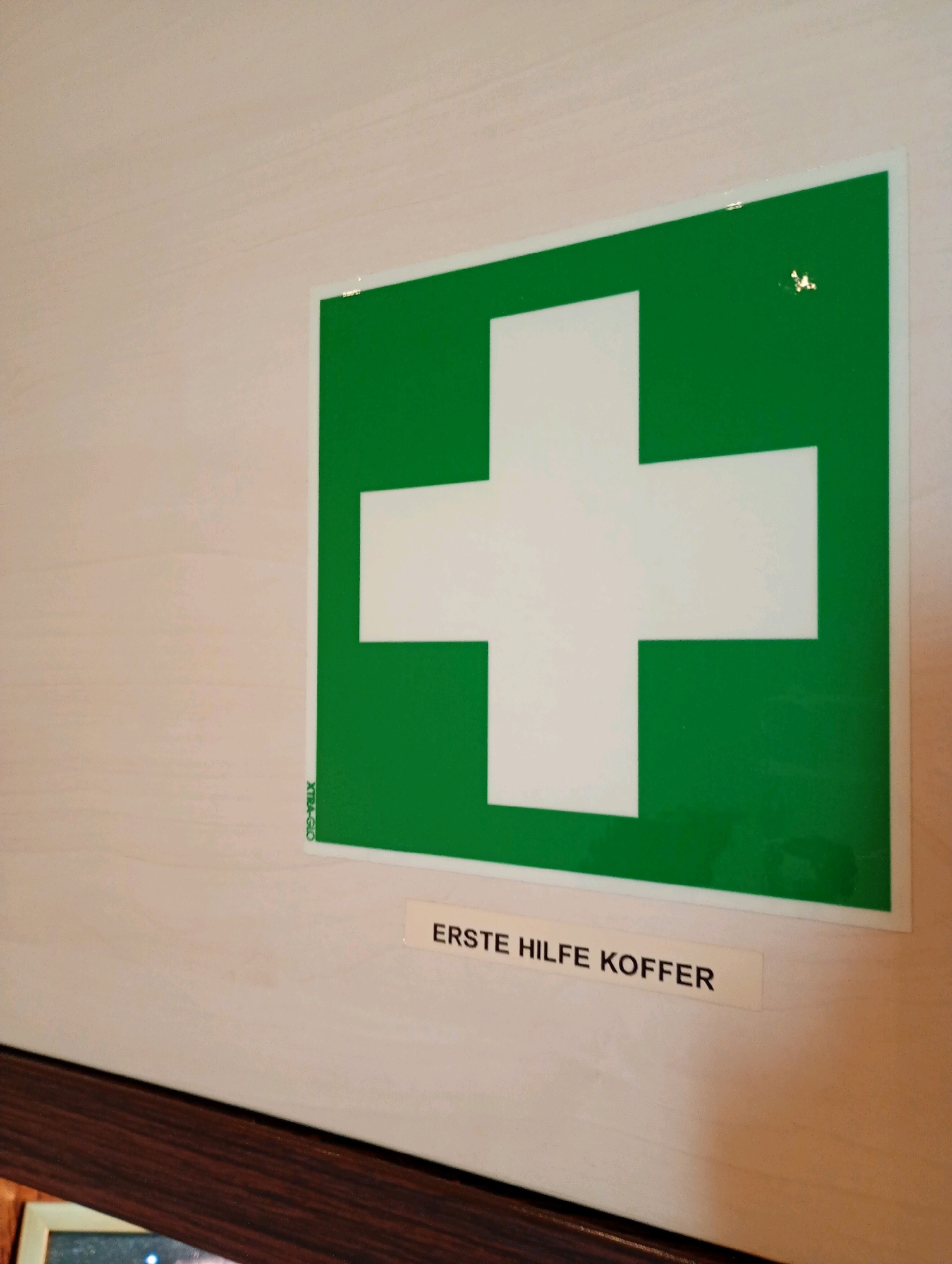 A first aid kit sign is displayed on a light-colored wall. The sign is green with a white cross in the center. Below the sign, a rectangular label reads "ERSTE HILFE KOFFER", which translates to "FIRST AID KIT". The context of Bad Sachsa, Germany is not discernable from the visual information provided.