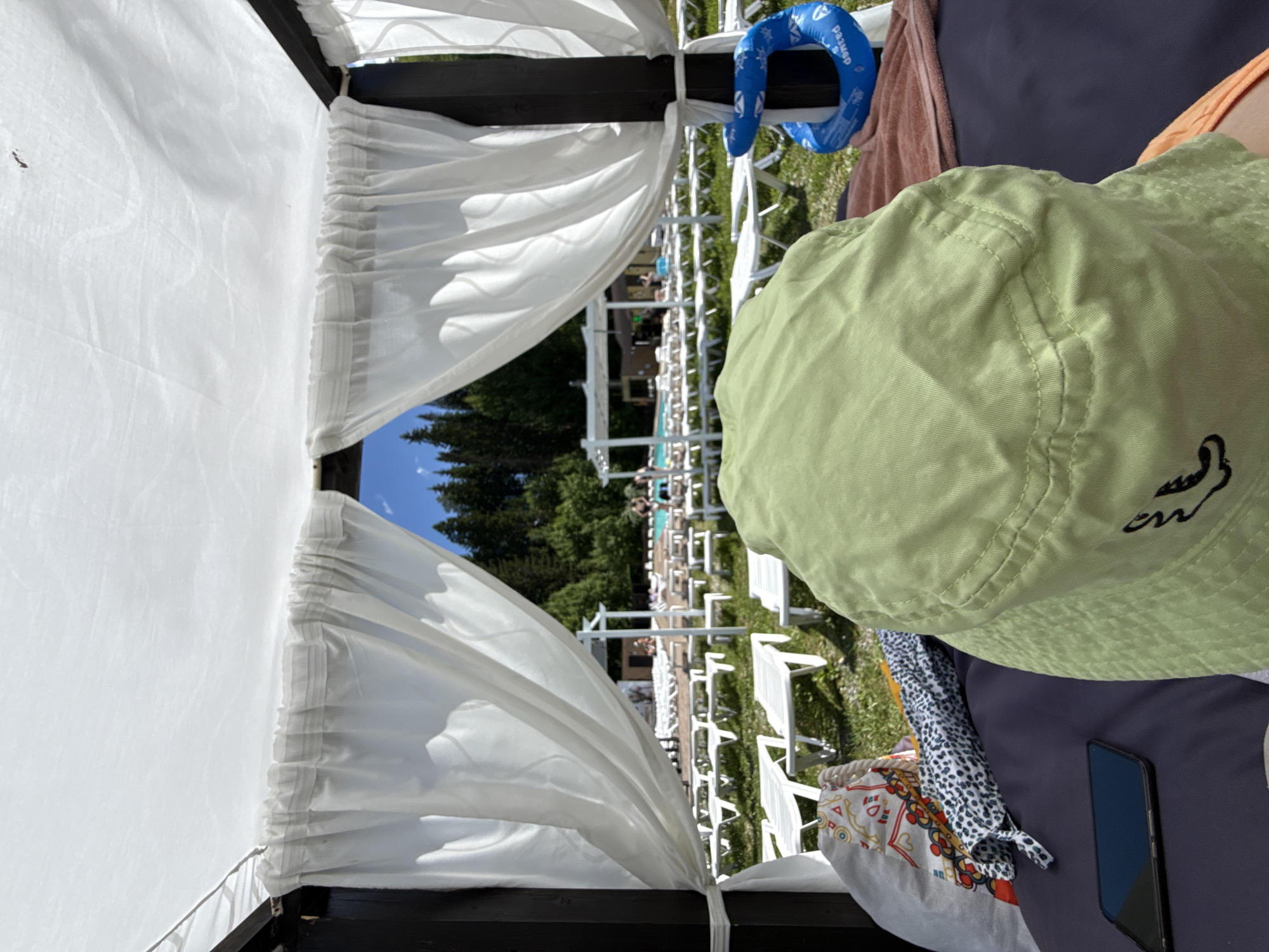 The image shows a personal perspective from under a canopy, looking out at an outdoor swimming pool area. In the foreground, a person wearing a light green bucket hat is seated on a dark surface, with a smartphone, a patterned fabric item, and a blue inflatable ring visible. The canopy above is white and draped with white sheer curtains. Beyond the canopy, rows of empty white lounge chairs are arranged on a grassy area. In the background, a pool with several people swimming is visible, along with trees and a clear blue sky. The location is identified as Nechkino, Russia.