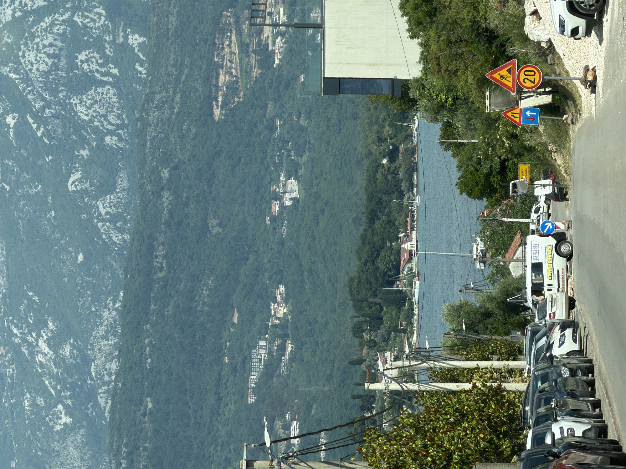 The image shows a view of Milovići, Montenegro, from an elevated position. A road lined with parked cars runs along the right side of the frame. The road features several traffic signs, including a construction warning sign, a speed limit sign indicating 20, and directional signs. A body of water stretches out beyond the road, dotted with boats and buildings along the shoreline. The landscape transitions into a densely forested area, with houses scattered throughout. In the distance, a mountain range is visible. A modern, minimalist building is located near the road. The scene appears to be taken on a clear, sunny day.
