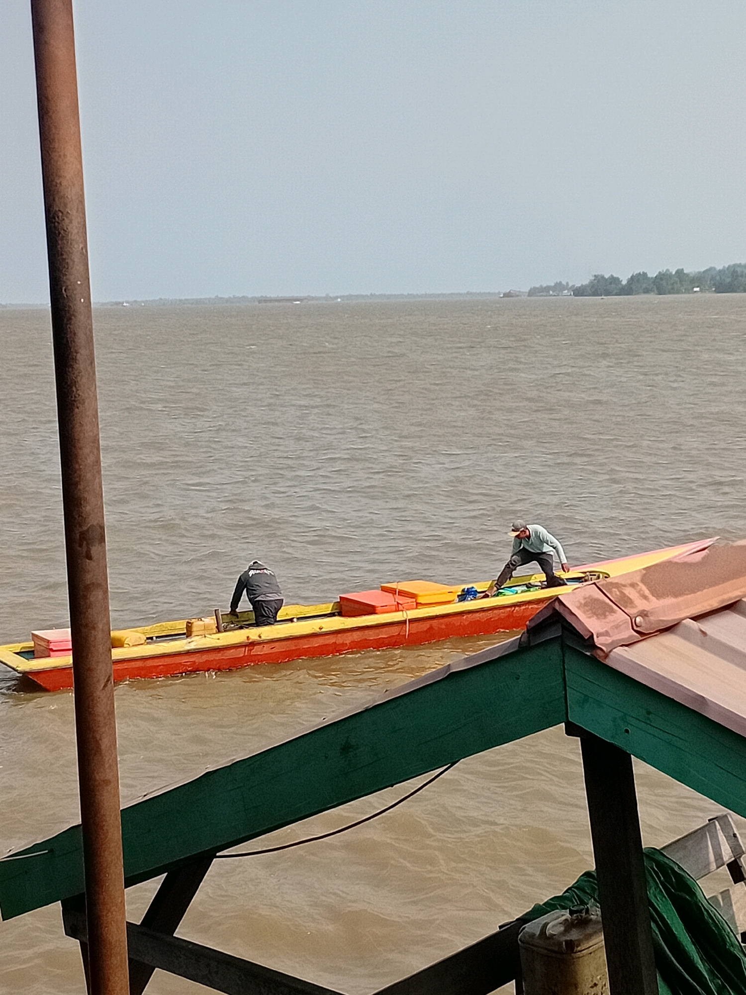 Two individuals are aboard a narrow, yellow and red boat on a body of water that appears to be a river or strait, under a hazy sky. One person, wearing a dark long-sleeved shirt and dark pants, is bent over near the front of the boat, partially obscured by a large brown pole in the foreground. The second person, wearing a light blue long-sleeved shirt and dark pants, with a grey cap, is crouched further back in the boat, appearing to be attending to items within the vessel. Several rectangular, brightly colored objects, possibly containers or buoys, are secured in the boat. The water is murky brown and shows moderate ripples. In the distance, a shoreline with sparse trees and some structures is visible. In the foreground, a portion of a structure with a green and pink roof, supported by dark wooden beams, extends into the frame. This scene is consistent with a riverine environment, potentially in a coastal area of Sungai Meriam, Indonesia, where such boats are commonly used for transport and commerce.