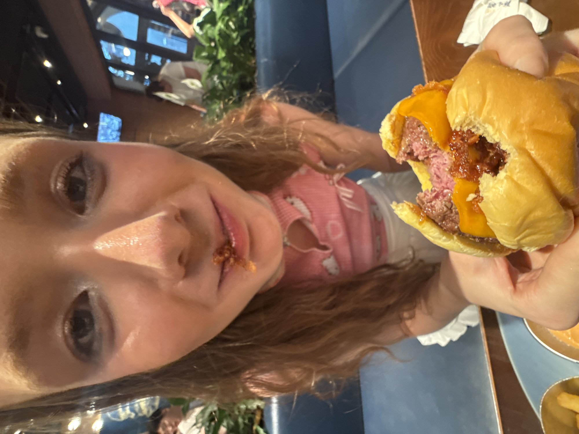 An extreme close-up shot shows a young person with reddish-blonde hair in a restaurant. The person is wearing a pink top with white cloud and heart designs. Their face is close to the camera, and there is food residue around their mouth, suggesting they have been eating. The person is holding a partially eaten hamburger with cheese and sauce. In the background, other patrons are visible, along with plants and restaurant decor. The setting appears to be a modern eatery in Pudong, China.