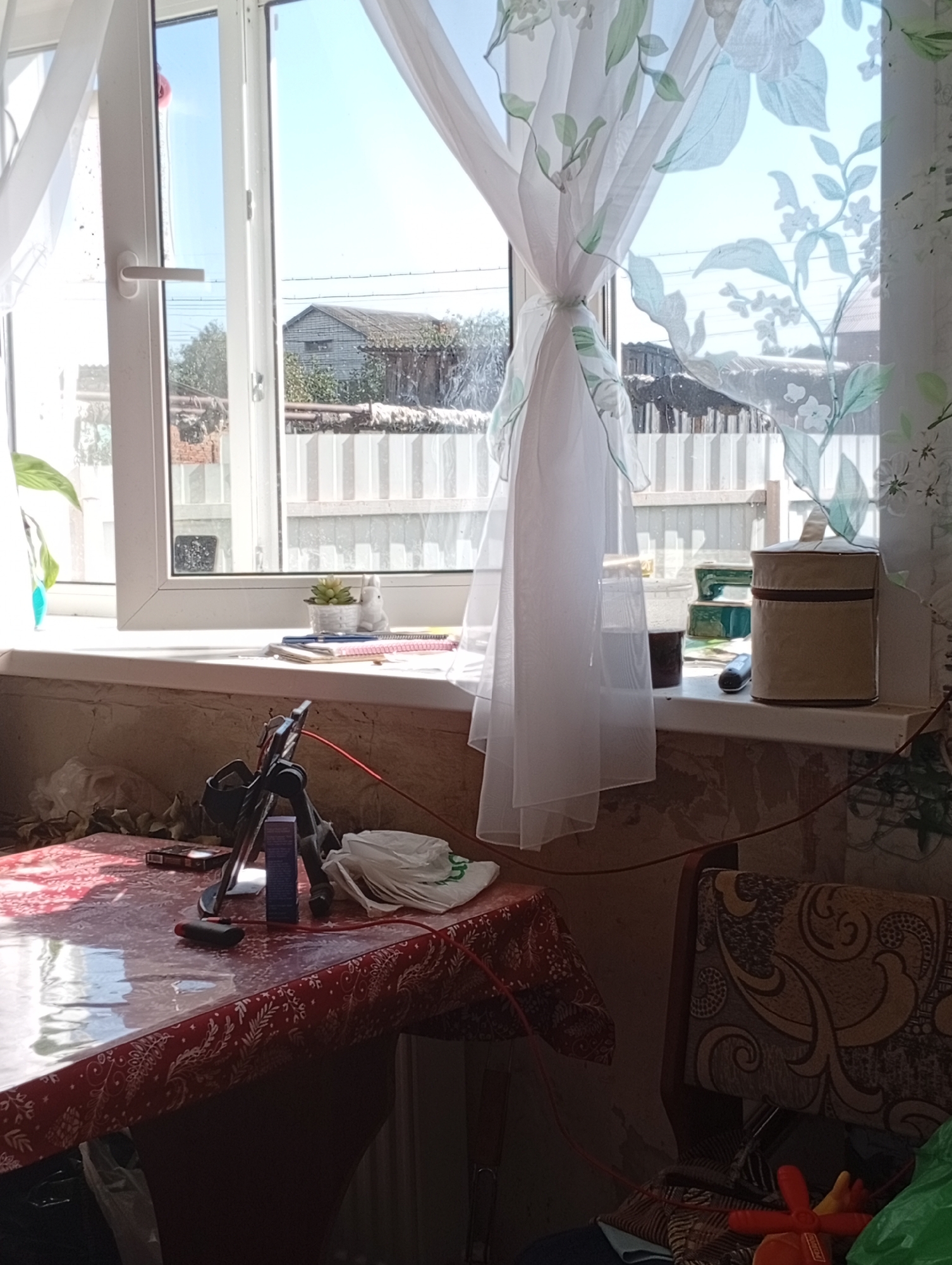 The image depicts an indoor scene, possibly a room, with a window in the background. The window looks out onto a residential area with houses and a fence. Sunlight streams in through the window, illuminating the room. In the foreground, there is a table covered with a red tablecloth. Various objects are scattered on the table, including a tripod with a phone, a small blue box, and some white folded items. To the right of the table, a chair with patterned upholstery is visible. The walls appear to be made of stone or concrete. The overall impression is one of a lived-in space with everyday objects.