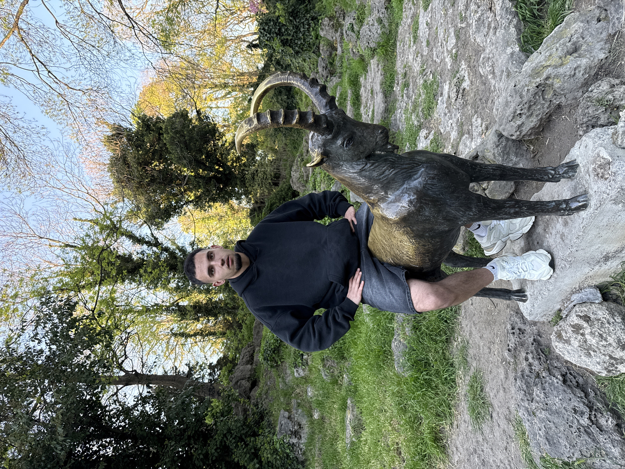 A man is sitting on a bronze statue of an ibex in a park-like setting. The man is fair-skinned with dark hair, wearing a black hooded sweatshirt and gray shorts. He is leaning to his left, with his right hand on the ibex's back. The ibex statue is positioned on a rocky outcrop surrounded by grass. The ground is covered with grass and rocks. Trees are visible in the background, and the sky is light blue. This is likely a park located in Varna, Bulgaria, known for such statues and natural landscapes.