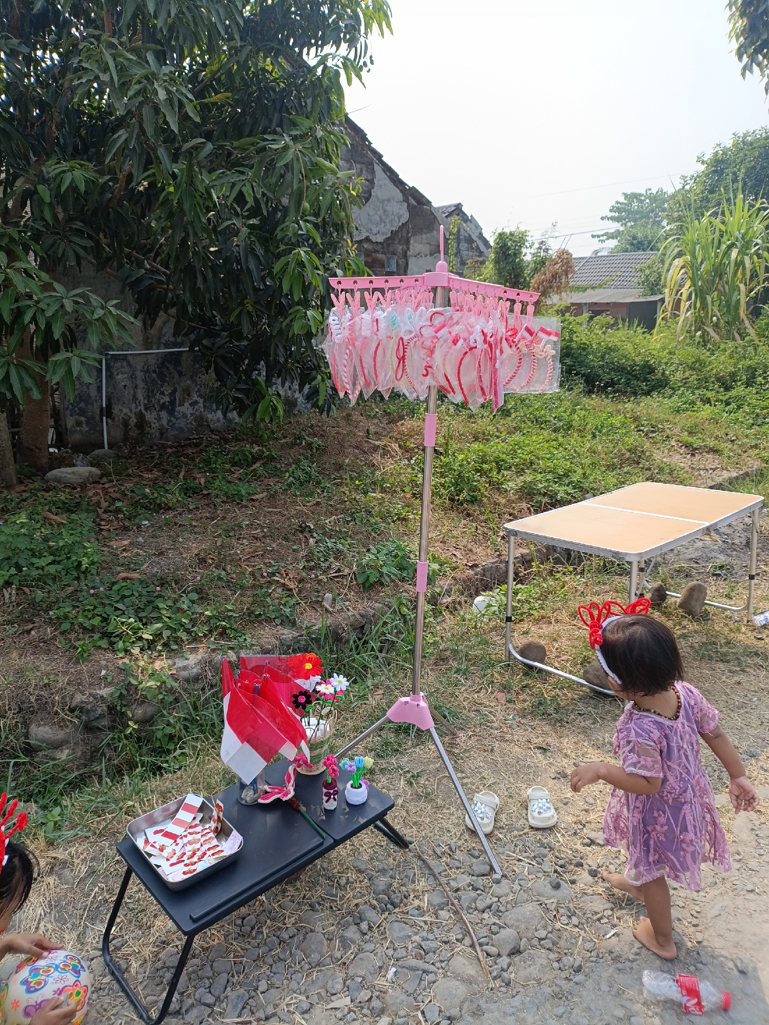 In Cariu, Indonesia, a young girl in a purple dress and a red headband stands in a yard. She is facing away from the camera, looking towards a small black table that holds several Indonesian flags, artificial flowers, and a tray of what appear to be small red and white objects. Nearby, a pink clothes drying rack holds many small, translucent items with red trim. A light-colored portable table stands to the right of the scene, with two large rocks beneath it. The ground is covered in gravel and dry vegetation. Behind the objects, a large tree with dense green foliage is visible, and in the background, a weathered building and more greenery can be seen under a bright sky. The girl is barefoot, and a pair of white sandals are on the ground near her. Another child's arm, holding a colorful ball, is visible in the lower left corner of the frame. A plastic water bottle is on the ground to the right.