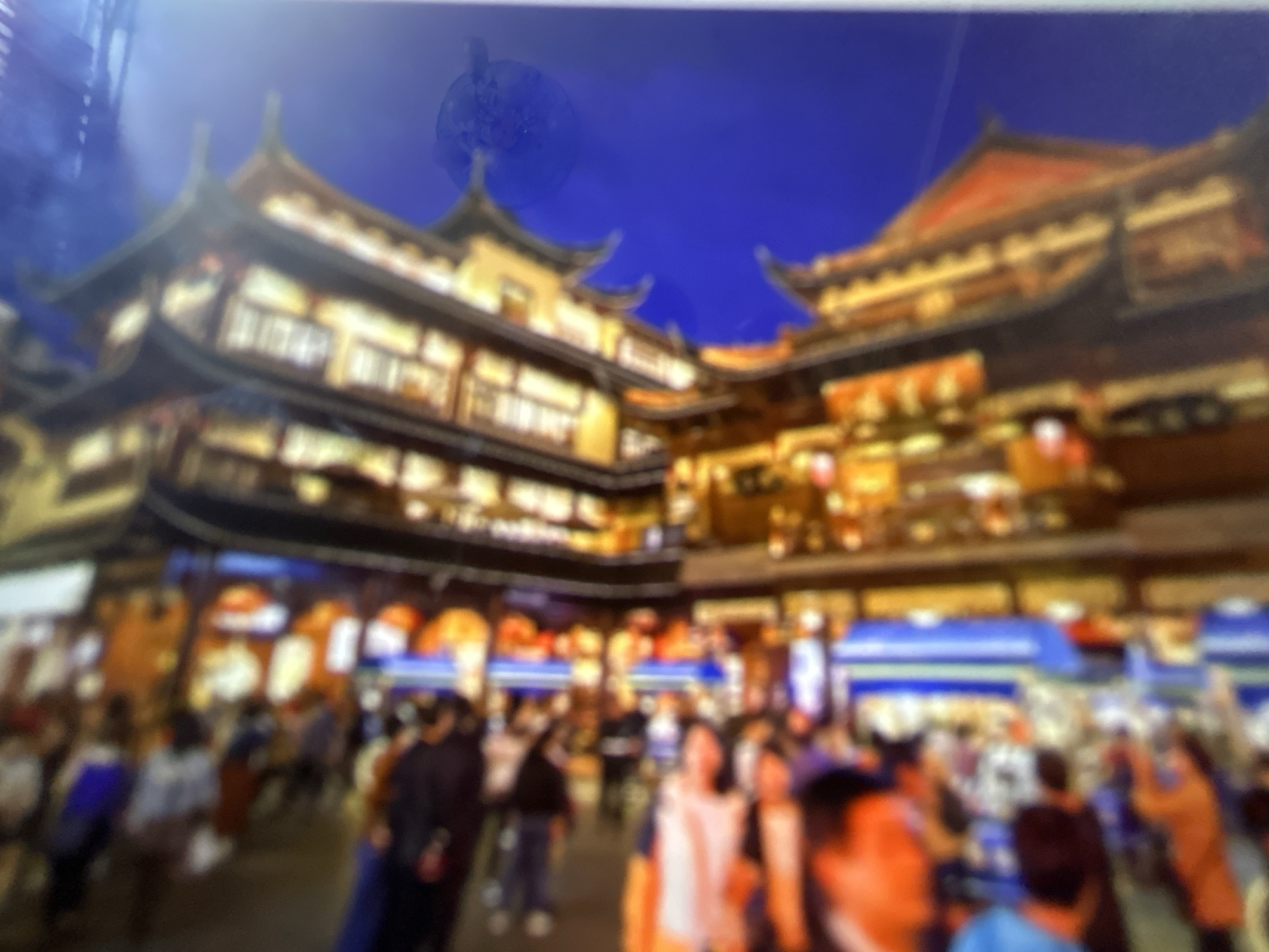 The image is a blurry, vertical photograph of a brightly lit, narrow street scene, likely at dusk or night. The architecture suggests a traditional or historical setting, possibly a marketplace or entertainment district. Buildings on either side of the street are adorned with numerous lights, creating a warm, golden glow. In the foreground, blurred figures of people are visible, some wearing brightly colored clothing. They appear to be walking or standing, engaged in some form of activity. The overall impression is one of a bustling, vibrant urban environment. The context provided about Gelendzhik, Russia, implies this scene might be a popular tourist area within the city.