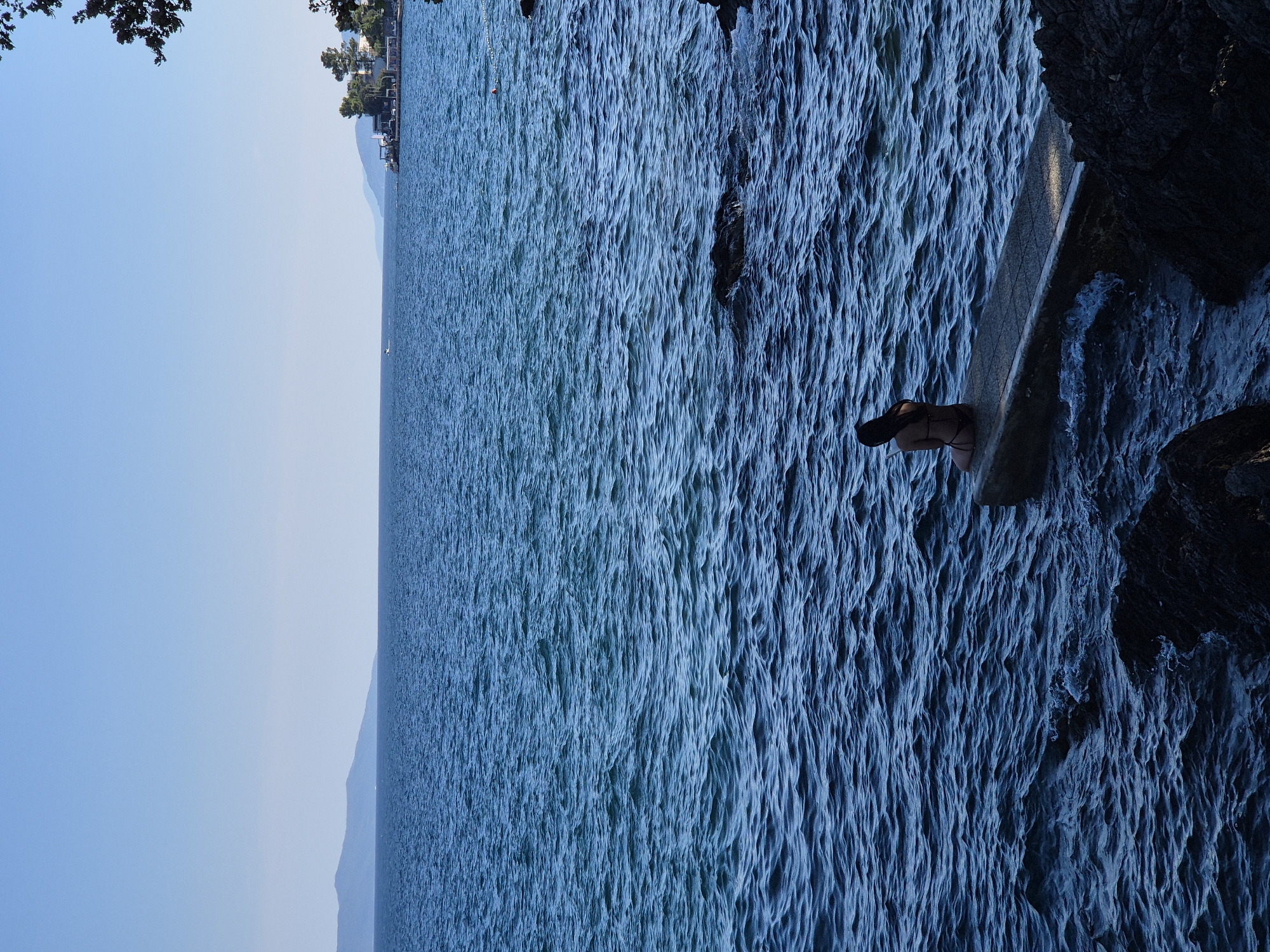 A lone figure, a woman in a bikini, sits on the edge of a concrete pier, facing the vast blue ocean. The water is a deep, vibrant blue, with gentle waves lapping against the rocks that surround the pier. In the distance, a hazy line of mountains borders the horizon, under a clear, pale blue sky. To the right, a small cluster of buildings and trees are visible along the coastline, and a few distant boats dot the seascape. The lighting suggests it is daytime, with the sun casting a slight shadow on the pier. The overall scene is serene and contemplative.