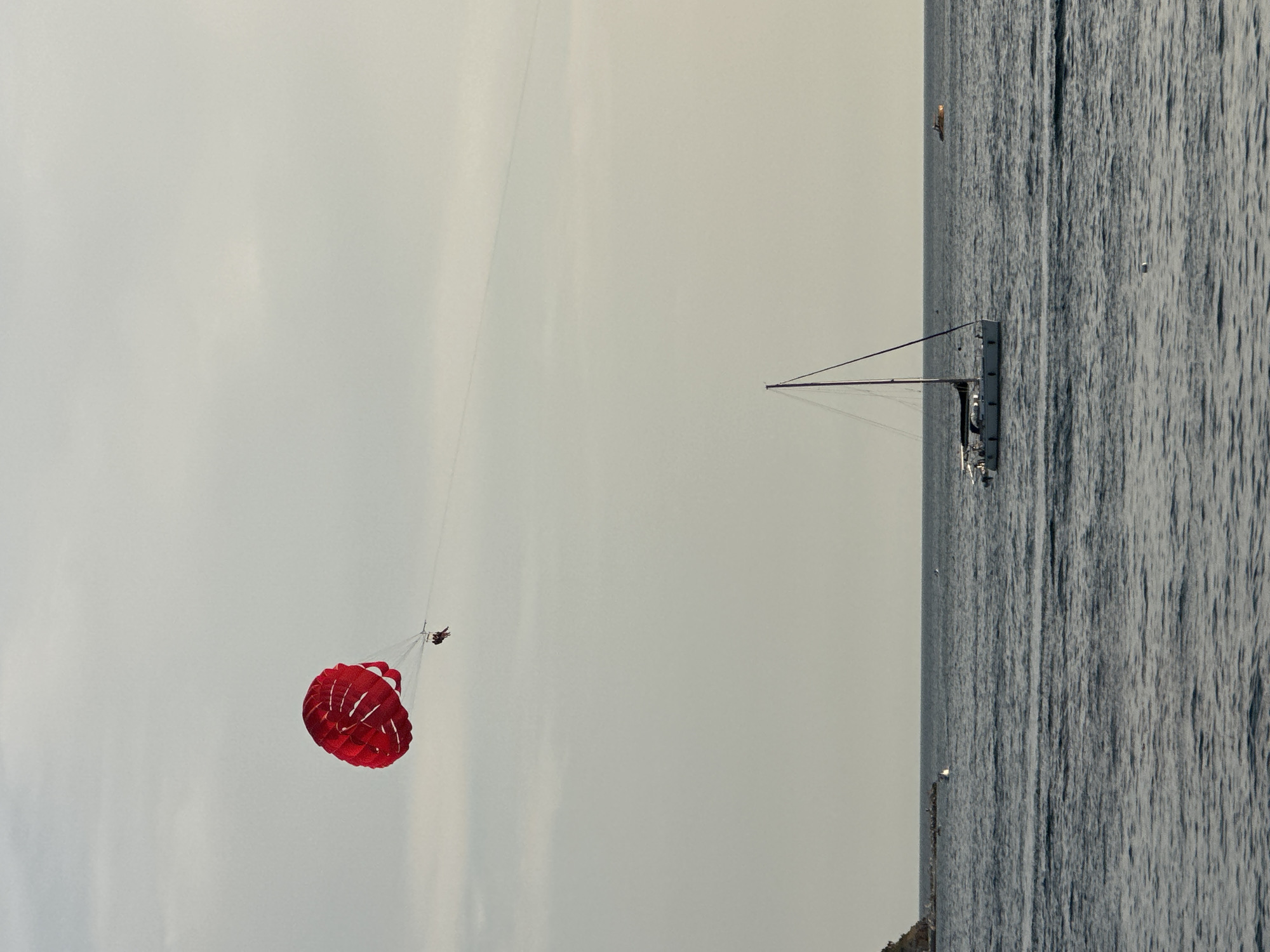 A parasailer, holding a red and white parachute, is being towed by a boat over the Adriatic Sea off the coast of Budva, Montenegro. The parasailer appears to be a person, and the parachute is open and catching the wind. A sailboat with a dark sail is anchored in the water, and a small yellow boat is visible in the distance. The sky is overcast, and the water is a deep blue. The scene suggests a recreational activity taking place in a coastal urban environment.