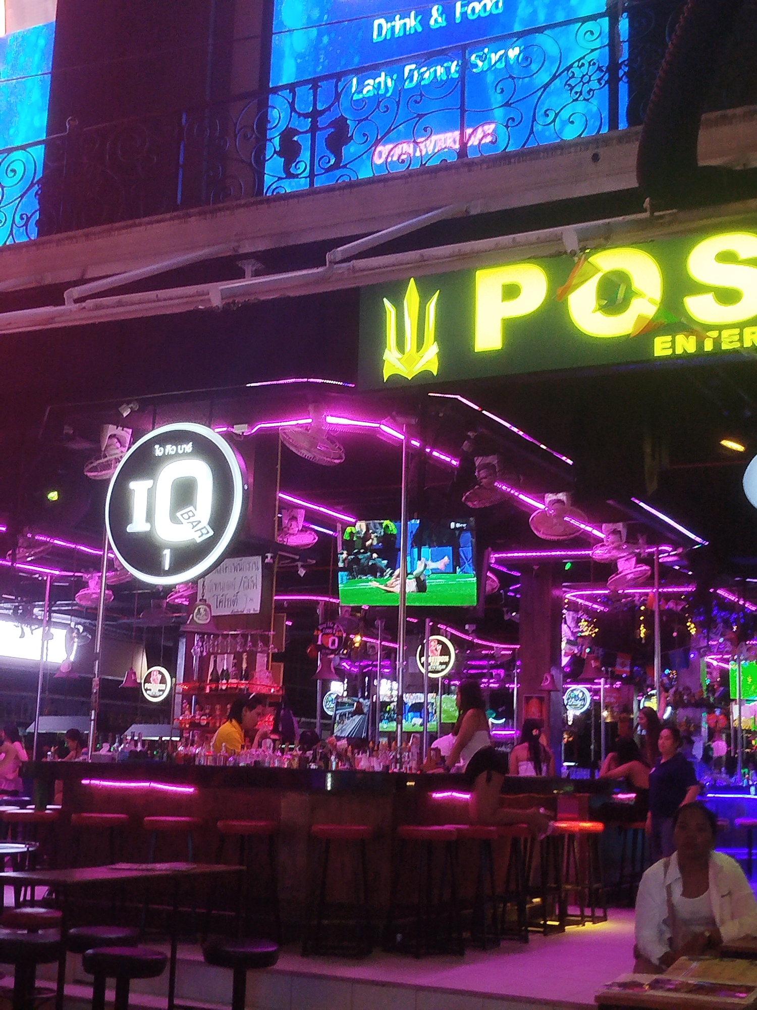 The image depicts the interior of a brightly lit bar with neon accents, likely a nightclub or entertainment venue, possibly in a tropical or Asian location given the signage and atmosphere. In the foreground, a woman in a white shirt and what appears to be a white bra is seated at a table, looking towards the camera. Behind her, a long bar area is visible, with several patrons sitting on stools. A bartender, wearing a yellow shirt, is visible behind the bar, attending to drinks. The bar is illuminated by pink neon lights. Above the bar, a large circular sign with "IQ BAR 1" written on it is prominently displayed. Further back, various screens are visible, some showing sports or other video content, and more neon lighting illuminates the space. A large, bright yellow sign that reads "POST ENTER" is affixed to the ceiling. The overall ambiance is that of a lively and active nightlife establishment.