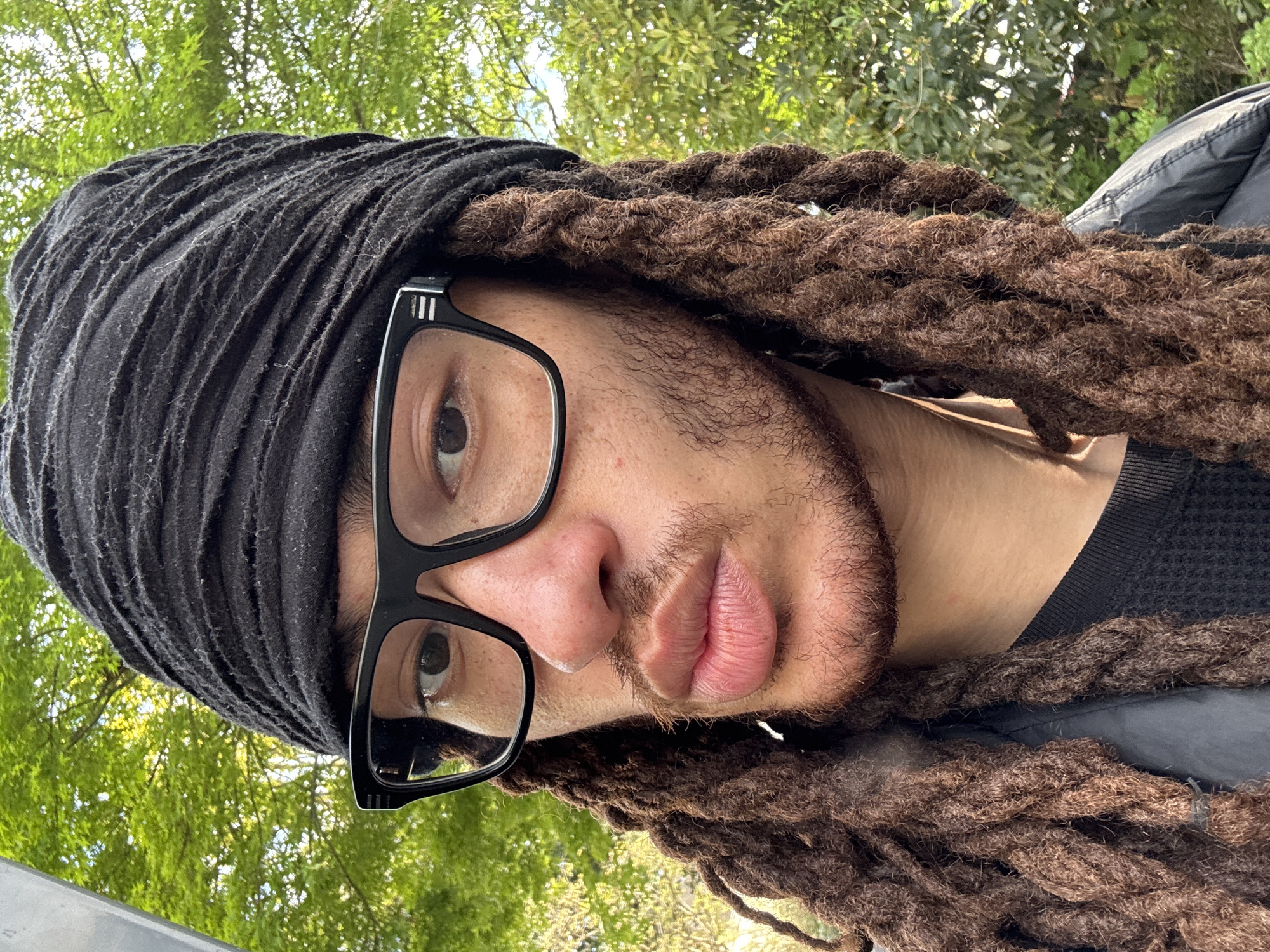 A close-up, tilted portrait shows a person with brown dreadlocks, some covered by a black head covering. The person wears black-framed glasses. Their face is visible from above the nose to below the mouth, showing facial hair. Green trees form the blurred background. The location is unknown and no location or city information is apparent within the image. Therefore, it is not possible to include any information about Les Ulis, France.