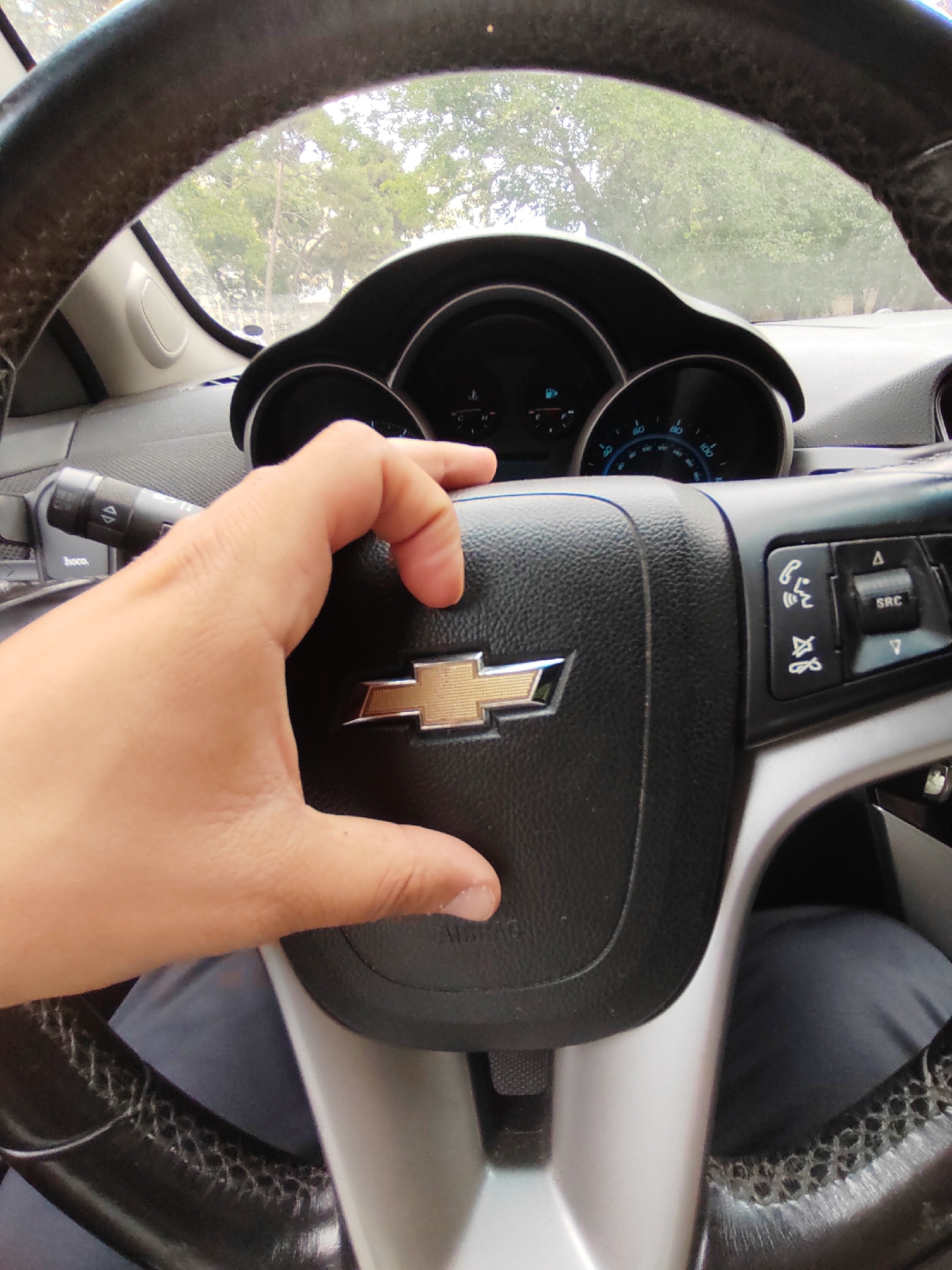 A first-person perspective shot captures a hand pressing the horn on a Chevrolet steering wheel. The steering wheel is black leather with a gold Chevrolet emblem in the center. To the right of the emblem are various buttons, including a call button, a microphone button, and a source button. The dashboard is visible behind the steering wheel, with a speedometer and tachometer. The interior of a car, possibly a Chevrolet Cruze, is shown. The scene is set outdoors with trees and a cloudy sky visible through the windshield. The image appears to have been taken in Baku, Azerbaijan, given the context of the surrounding environment.
