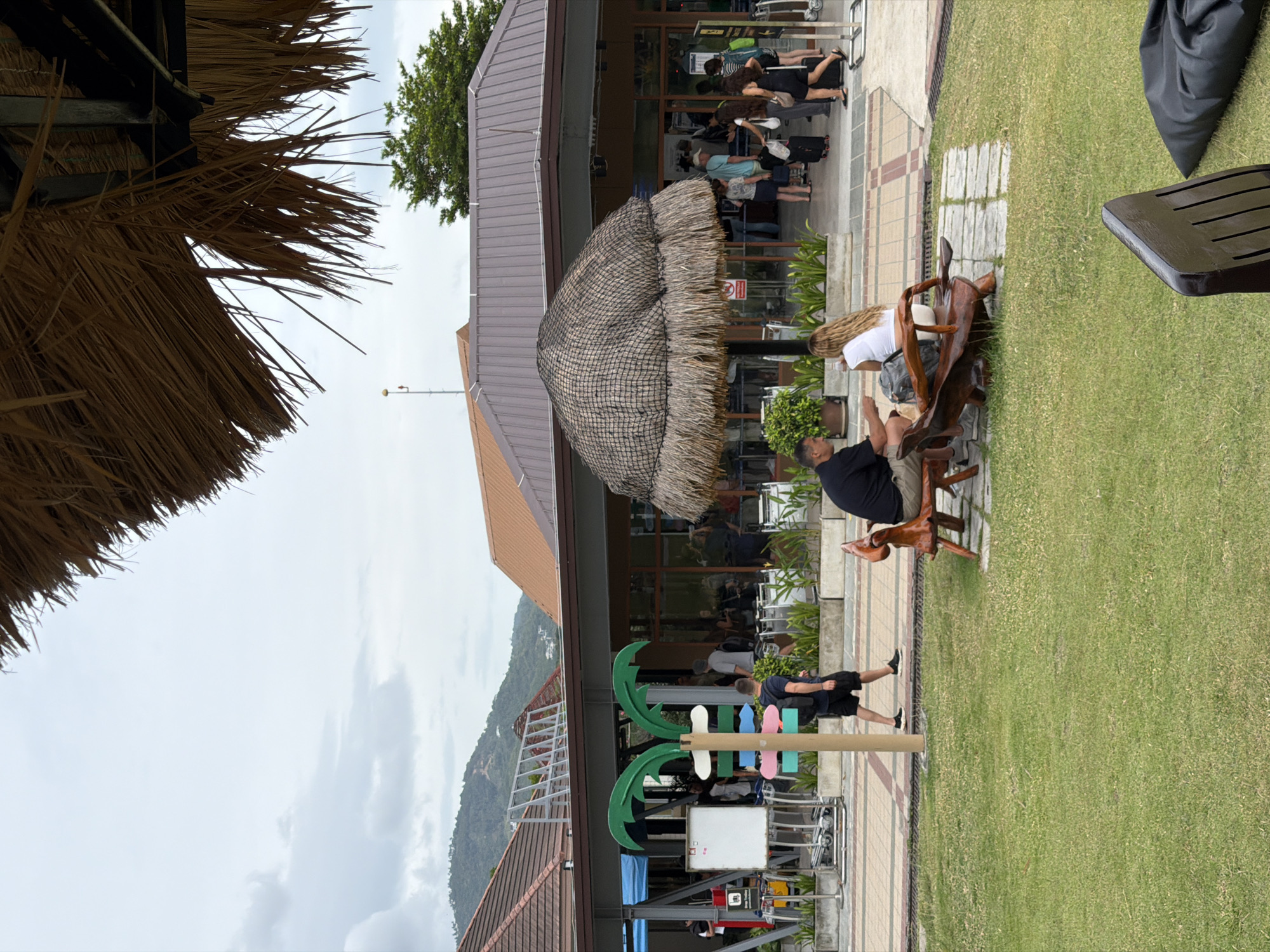 In Ko Samui District, Thailand, this image depicts an outdoor waiting area at what appears to be a ferry terminal or transportation hub. In the foreground, a person with dark hair is seated on a wooden bench on the left, facing away from the camera. To their right, a woman with long blonde hair is seated on a uniquely shaped wooden bench, facing left. Both individuals are dressed casually. Behind them, a pathway runs parallel to a building with a brown, sloped roof. Several people are walking and standing in front of the building, some with luggage, suggesting travel. A tiki-style thatched umbrella stands in front of the building. To the left, a colorful signpost with palm tree-like branches and directional arrows stands near a whiteboard. The ground is a mix of paved areas and a green lawn. In the background, lush green hills rise under a cloudy sky. A man wearing a backpack and dark shorts walks away from the camera on the paved path.