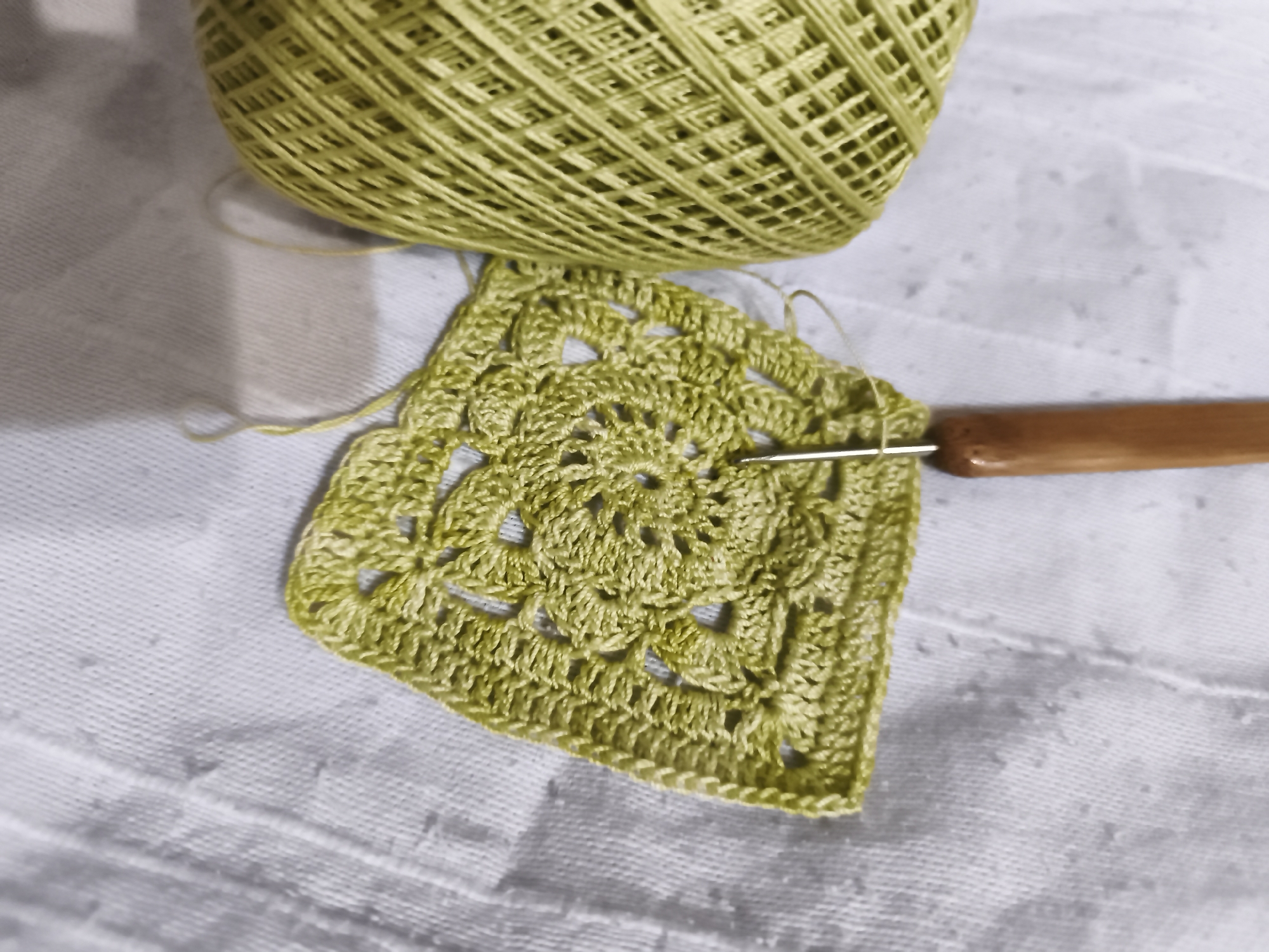 The image displays a close-up, high-angle shot of a ball of light green yarn and a partially crocheted square piece of fabric in the same color. A crochet hook with a wooden handle is inserted into the crocheted fabric, indicating that it is currently being worked on. The fabric exhibits a delicate, lacy pattern. The items are placed on a textured surface that appears to be a light-colored fabric, possibly a tablecloth or bedding, with visible stitching lines. The background is a lighter shade of the same fabric, creating a soft and muted backdrop. The lighting is soft and diffused, casting subtle shadows. There are no people present in the image, and no specific city or location is identifiable from the visual cues.