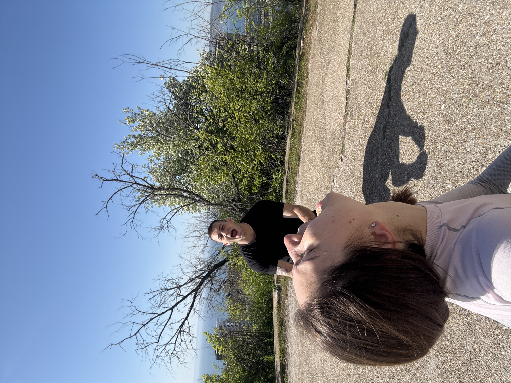 A sunny outdoor scene shows two people on a concrete surface. A woman with short brown hair and wearing a light-colored long-sleeved shirt is positioned towards the bottom of the frame. Behind her and slightly to the left, a man wearing a black t-shirt is visible. He is looking at the camera with his mouth open. The background includes a tree with green leaves, bare branches, and a clear blue sky. The shadow of a person, possibly taking the picture, is cast on the concrete. A body of water is visible in the distance, suggesting a location near a shore. The specific location within the city of Unknown is not discernible from the image.