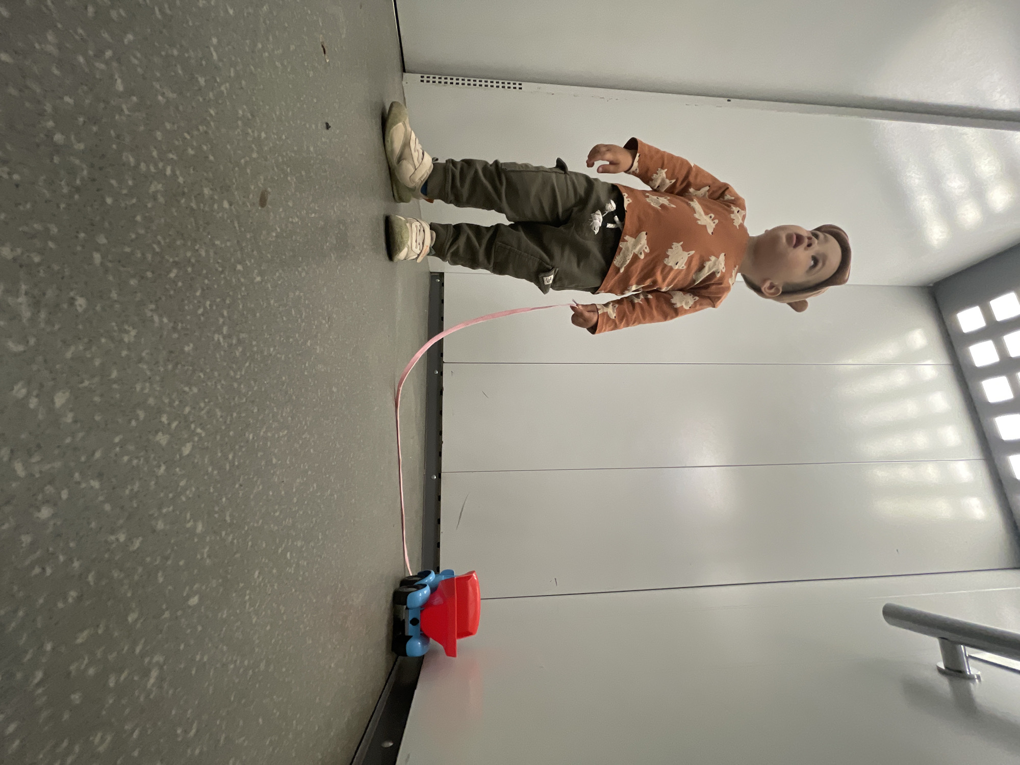 The scene is set in an indoor environment within Izhevsk, Russia, specifically a confined space such as an elevator or a small room characterized by white paneled walls and a grey, speckled floor. A young child, likely a toddler, stands slightly left of the frame's center, facing partially right and looking upwards with a slightly open mouth. The child wears an orange long-sleeved shirt adorned with light-colored, cartoonish dog patterns, dark olive-green cargo pants with pockets on the thighs, and light beige sneakers with Velcro straps. A brown cap, featuring two small, rounded ear-like protrusions, is worn on the child's head.
The child's right hand holds a thin pink strap or ribbon, which extends across the floor to the right. This strap is connected to a small toy dump truck positioned in the lower right corner of the frame, near the wall. The toy truck has a blue base and a red dump bed.
The walls are predominantly white, smooth, and composed of large panels with visible vertical seams. Reflections of overhead light sources are present on these wall surfaces. A metallic, cylindrical handrail is mounted horizontally on the upper right wall. The ceiling, visible in the upper portion of the image, contains multiple square, recessed light fixtures emitting bright white light. The floor is a uniform grey with a distinctive pattern of light grey and white flecks, consistent with a terrazzo or composite material. Minor dark debris is visible on the floor. The perspective is low-angle, approximately at the child's knee height, looking upwards towards the child and the ceiling.