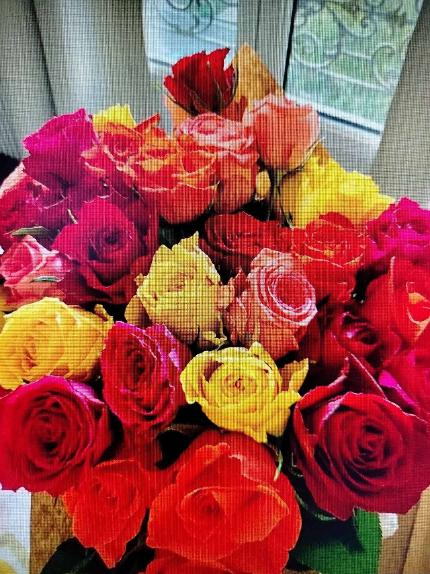 A close-up shot captures a vibrant bouquet of roses. The roses are a mix of deep reds, bright yellows, and fiery oranges, with some hints of pink. They are arranged densely, with some blooms fully open and others still partially closed, showcasing their intricate petal structures. The lighting suggests an indoor setting, possibly near a window, as indicated by the soft illumination and the blurry background that appears to be a curtain or blinds. There are no people or animals present in the image, and the focus is solely on the floral arrangement. The image does not provide any information about the location in Yashkino, Russia.