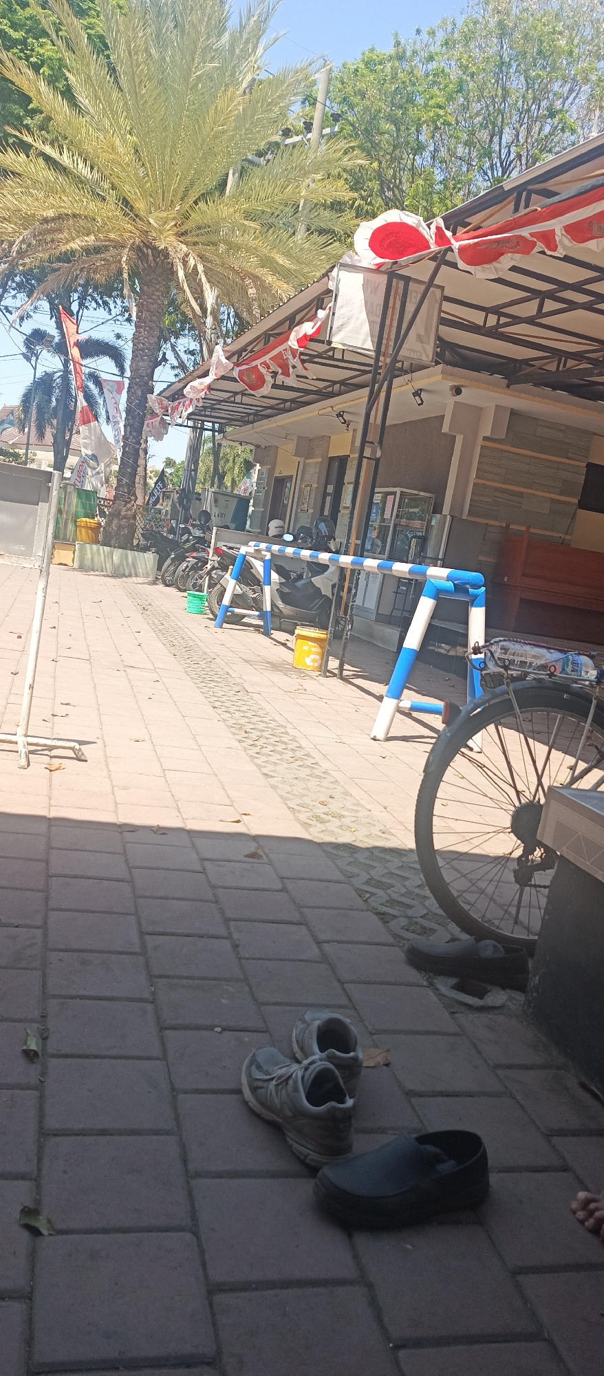 The image captures an outdoor scene on a sunny day, likely in a public area in Situbondo, Indonesia. A prominent date palm tree stands tall on the left, its fronds spread widely, casting a shadow over the tiled walkway. In the background, a low-rise building with a metal awning decorated with red and white banners is visible. Several motorcycles are parked in front of the building, suggesting a transportation hub or a place frequented by people arriving on motorbikes. A blue and white striped barrier is positioned on the walkway, separating the area. On the foreground of the tiled path, a pair of grey sneakers and a pair of black slip-on shoes are placed on the ground. A bicycle wheel is also visible on the right side of the image. The ground is paved with rectangular tiles. The sky is clear and blue.