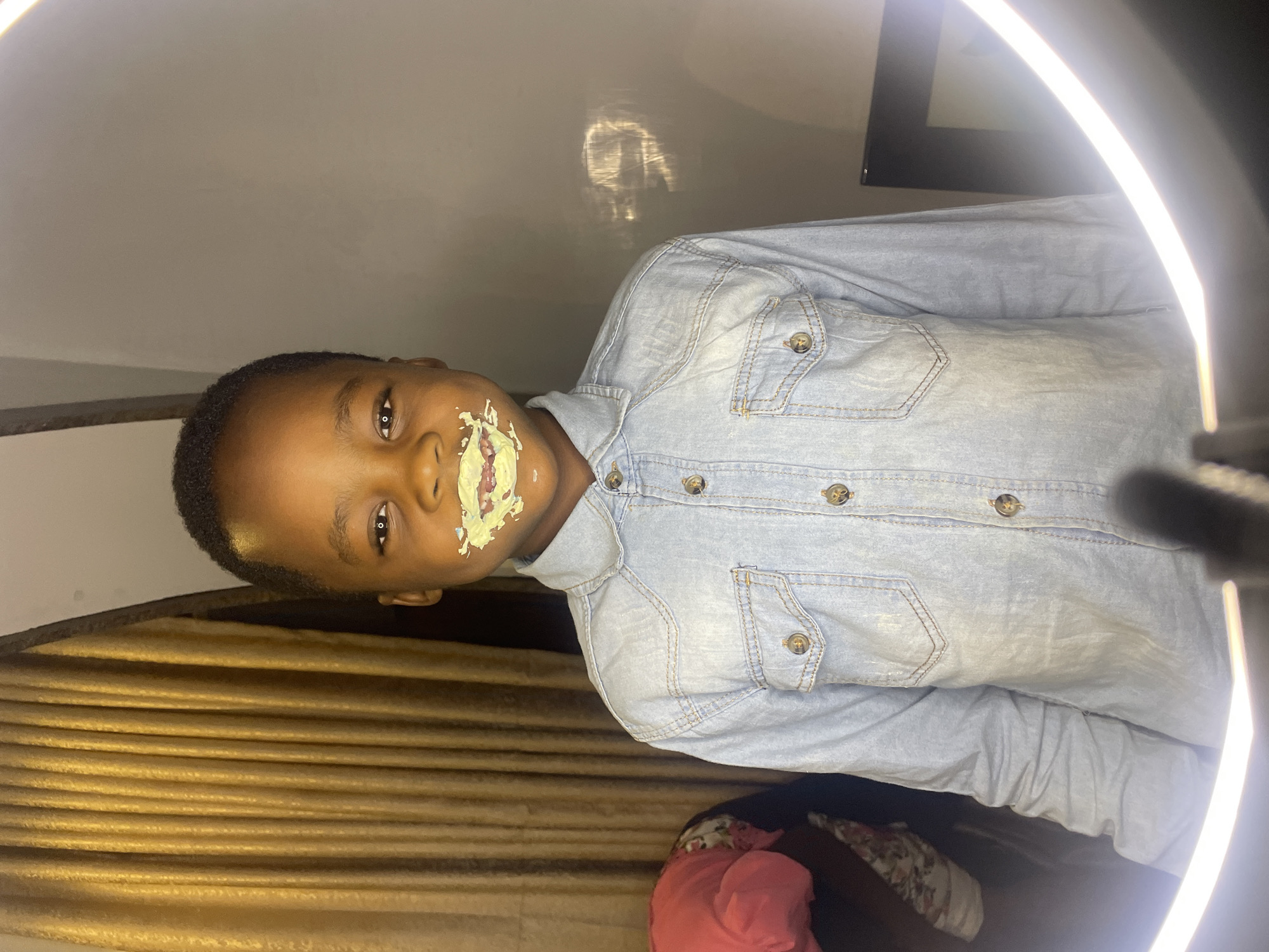 A young Black boy with short, dark hair is positioned in the center of the frame. He is wearing a light blue denim shirt. There is what appears to be cream or cake frosting smeared around his mouth.
The background includes golden-brown curtains, a partially visible black frame on the wall, and a bright ring light to the right of the frame. Due to the lack of identifiable landmarks or cultural details, it's not possible to confirm the location as Jos, Nigeria, from the image alone. However, if that information is provided as a known fact, then the setting is likely indoors within a home or studio in Jos, Nigeria.
