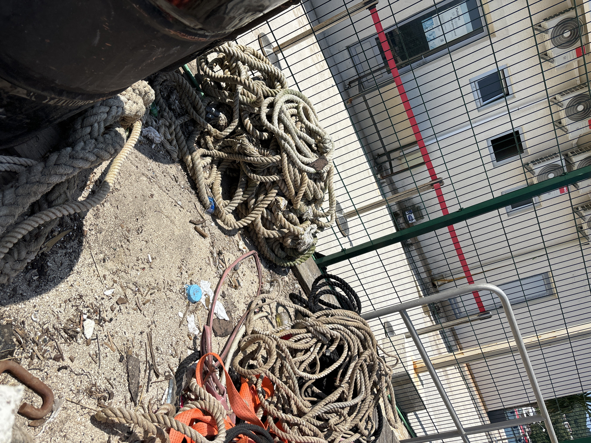 In Budva, Montenegro, at a sunlit outdoor location behind a wire mesh fence, several coils of thick, weathered rope are piled on sandy ground. The ropes vary in color from tan to dark brown and black. Some of the ropes are interwoven with orange webbing straps. A large, dark cylindrical object, possibly a buoy or a storage drum, is partially visible on the left side of the frame. Behind the fence, a light-colored building with multiple air conditioning units mounted on its exterior wall is visible. A red horizontal stripe is present on the building's facade, and two dark windows are visible. The ground is covered in sand, small debris, and some scattered pieces of plastic. A metal railing runs along the fence line in the middle ground. The scene appears to be a service or storage area, possibly associated with maritime activities given the presence of the ropes.